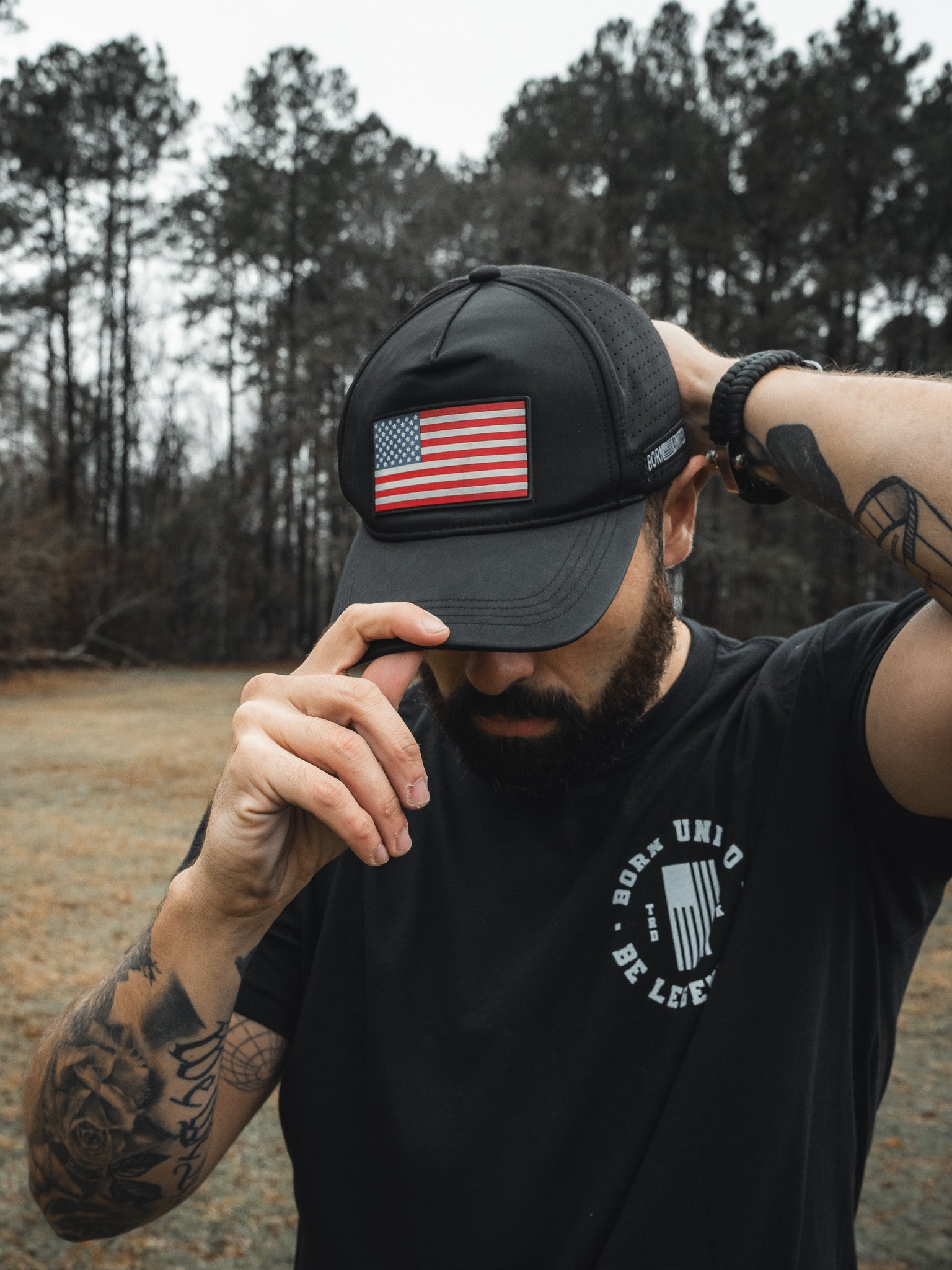 A bearded man, wearing the Born United Premium Flag Snapback - RW&B from Born United Private Label adjusts it outdoors. He sports a black T-shirt with white text and has tattoos on his arm, set against trees and a grassy field backdrop.