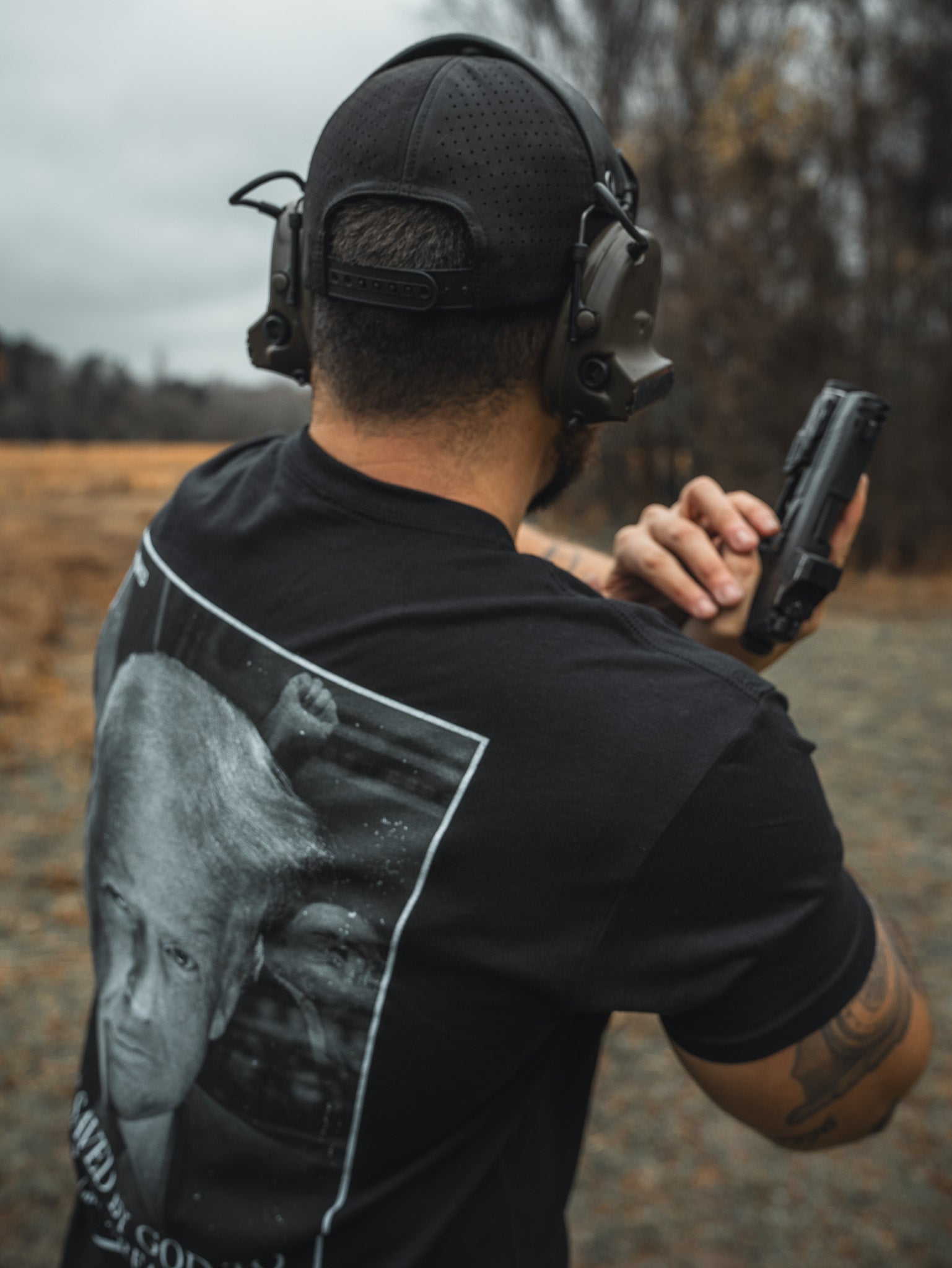 A person wearing a Born United Premium Flag Snapback - RW&B by Born United Private Label and a headset is at an outdoor shooting range, holding a handgun. Their shirt features a printed image against trees and an overcast sky, creating a distinctly American atmosphere.