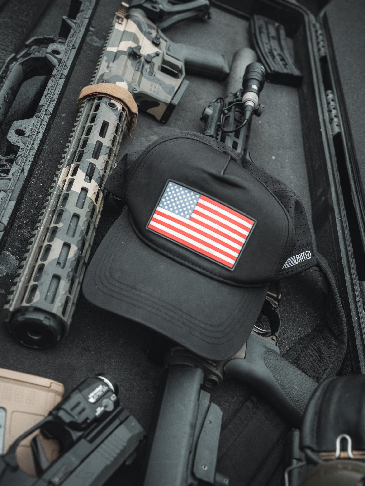 A Born United Premium Flag Snapback - RW&B sits atop a sleek black case filled with firearms and accessories, including rifles and a handgun with camouflage accents. This display from Born United Private Label embodies rugged patriotism.