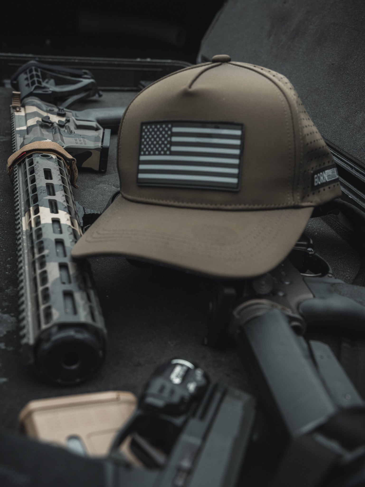 An olive Born United Premium Flag Snapback featuring a black American flag rests on a tactical rifle with attachments inside a vehicle with a dark, textured interior, creating a rugged vibe.
