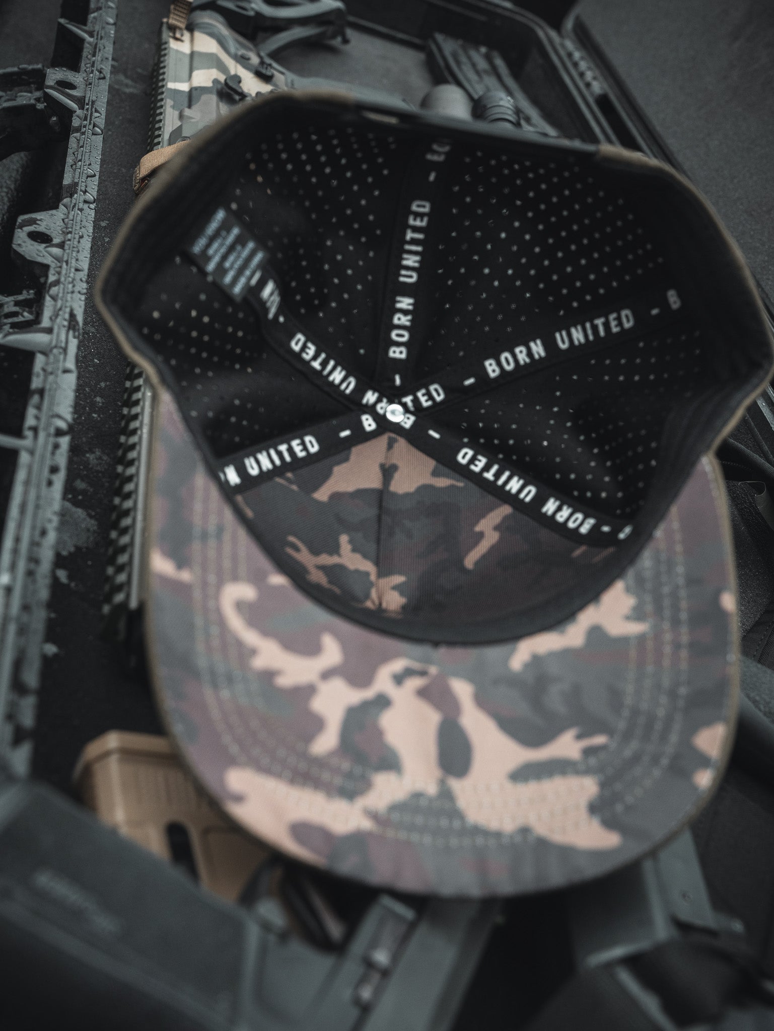 The Born United Premium Flag Snapback - Olive, a handmade camo cap featuring BORN UNITED text inside, rests on a rifle case alongside tactical accessories, embodying a military-inspired theme.