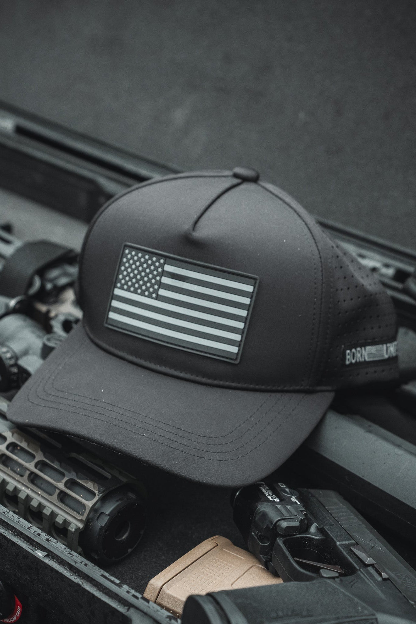 A Born United Premium Flag Snapback - Blackout from the Born United Private Label rests on tactical gear, alongside a rifle with a textured handguard and optical sight, in what appears to be a military or outdoor setting.