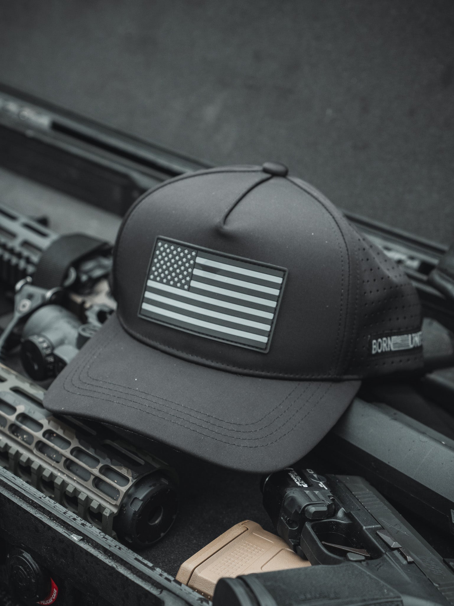 A Born United Premium Flag Snapback - Blackout from the Born United Private Label rests on tactical gear, alongside a rifle with a textured handguard and optical sight, in what appears to be a military or outdoor setting.