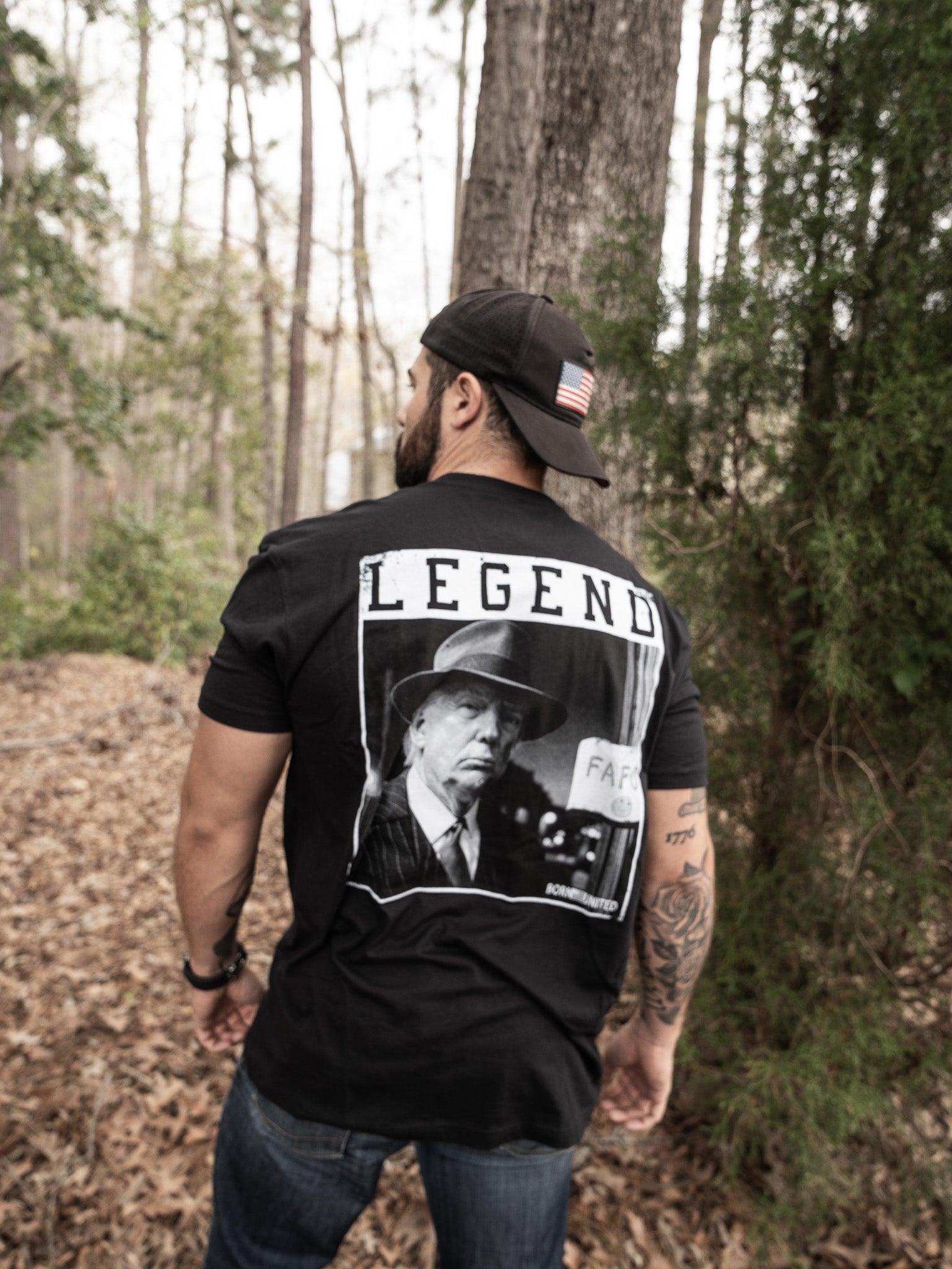 In a forest surrounded by trees and fallen leaves, a bearded, tattooed man stands wearing a black Born United Private Label cap featuring a USA flag and the Legend FAFO T-shirt with an image of a stern-looking man.