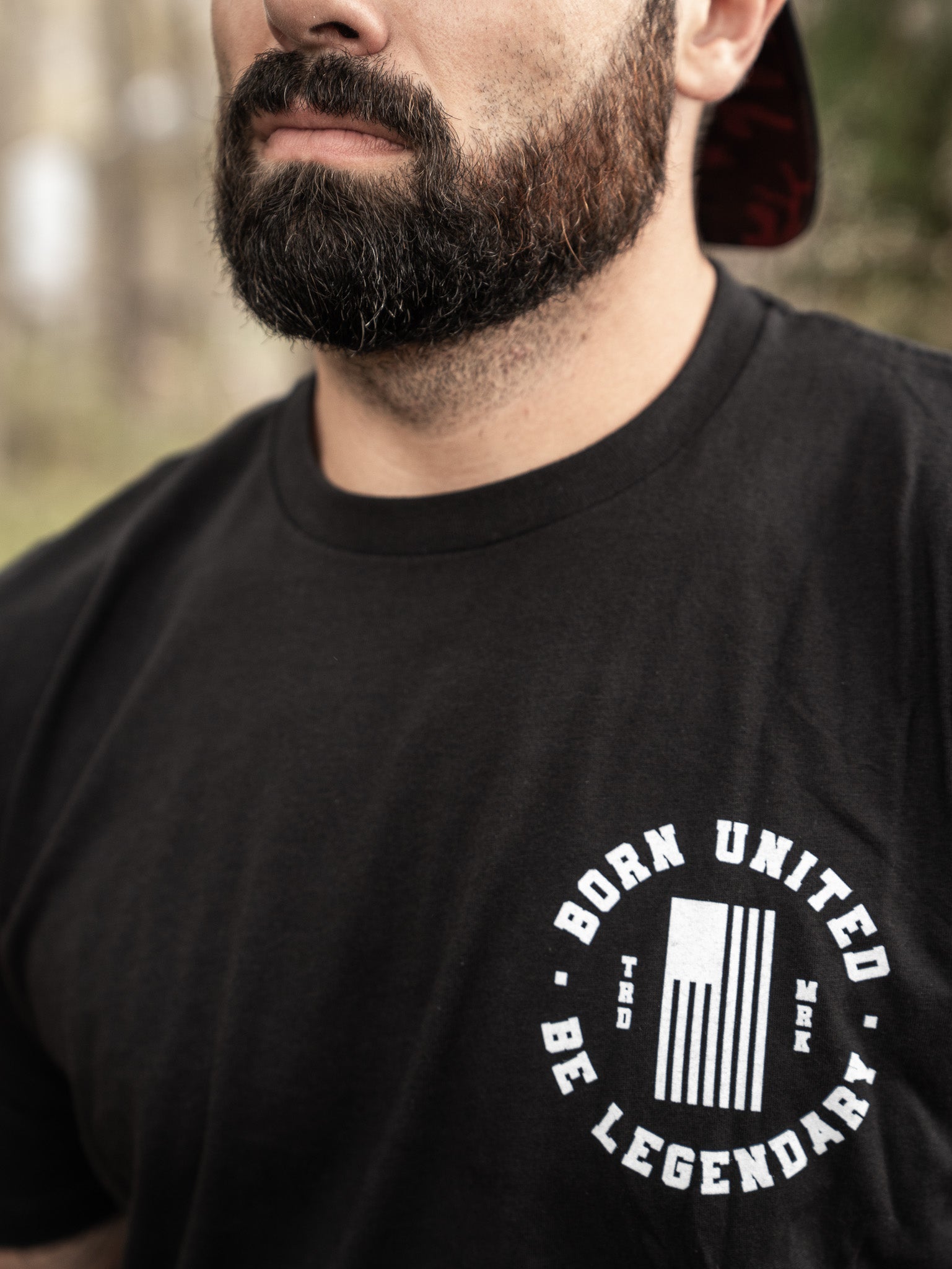 A bearded individual dons a black Born United Private Label t-shirt, BORN UNITED, BE LEGENDARY encircling a stylized flag logo. They stand outdoors against a blurred forest backdrop. Product: Legend FAFO.