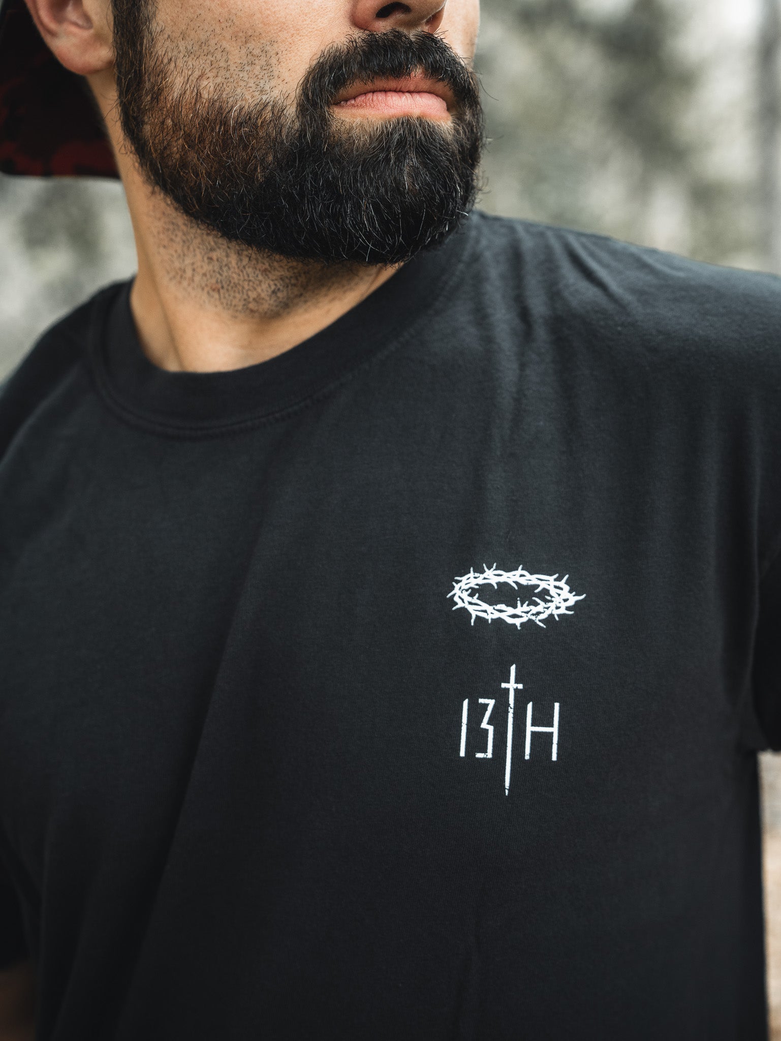 A bearded man stands out against a blurred background wearing the Jesus Saves - Black Tee from 13th, featuring a crown of thorns graphic and 13TH text in white, subtly echoing themes of everlasting life.