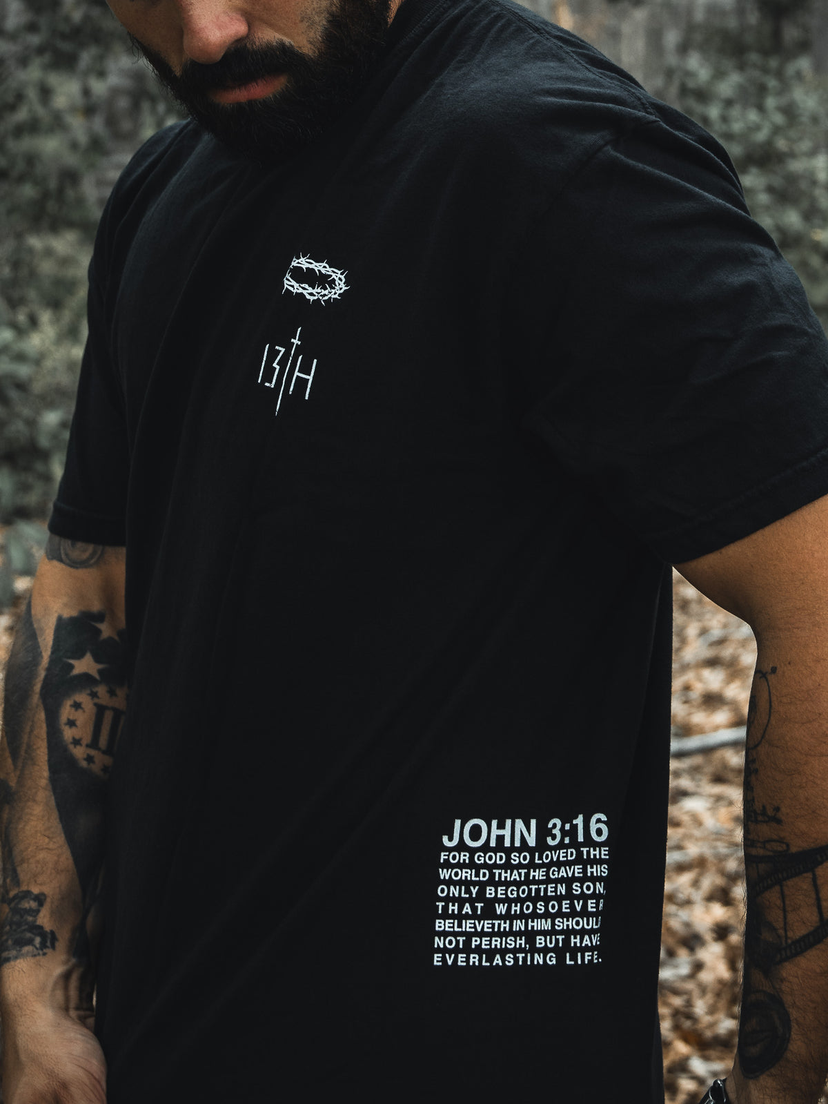 A tattooed individual wears the Jesus Saves - Black Tee from 13th, featuring a minimalist design with JOHN 3:16 and a quote about Gods love and His begotten son offering everlasting life. The background showcases blurred foliage.