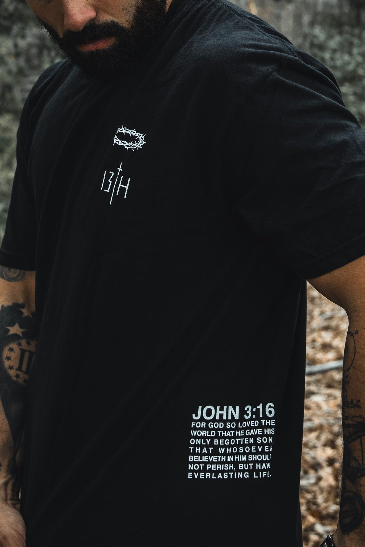 A tattooed individual wears the Jesus Saves - Black Tee from 13th, featuring a minimalist design with JOHN 3:16 and a quote about Gods love and His begotten son offering everlasting life. The background showcases blurred foliage.