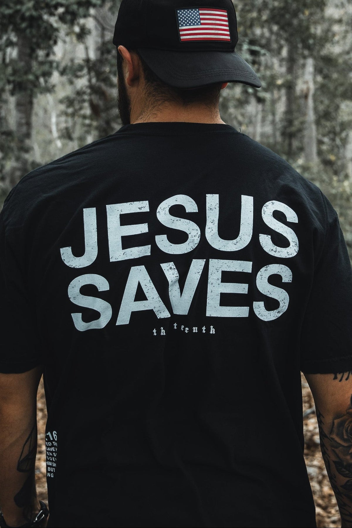 In a wooded area, a man dons the Jesus Saves - Black Tee by 13th and a black cap with an American flag. His tattooed arms suggest his deep faith, possibly inspired by John 3:16s promise of everlasting life through the begotten son.
