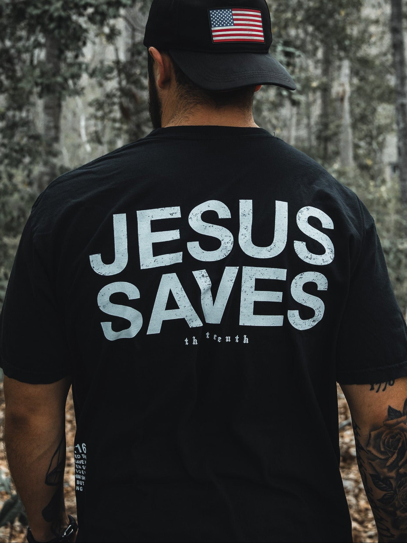 In a wooded area, a man dons the Jesus Saves - Black Tee by 13th and a black cap with an American flag. His tattooed arms suggest his deep faith, possibly inspired by John 3:16s promise of everlasting life through the begotten son.