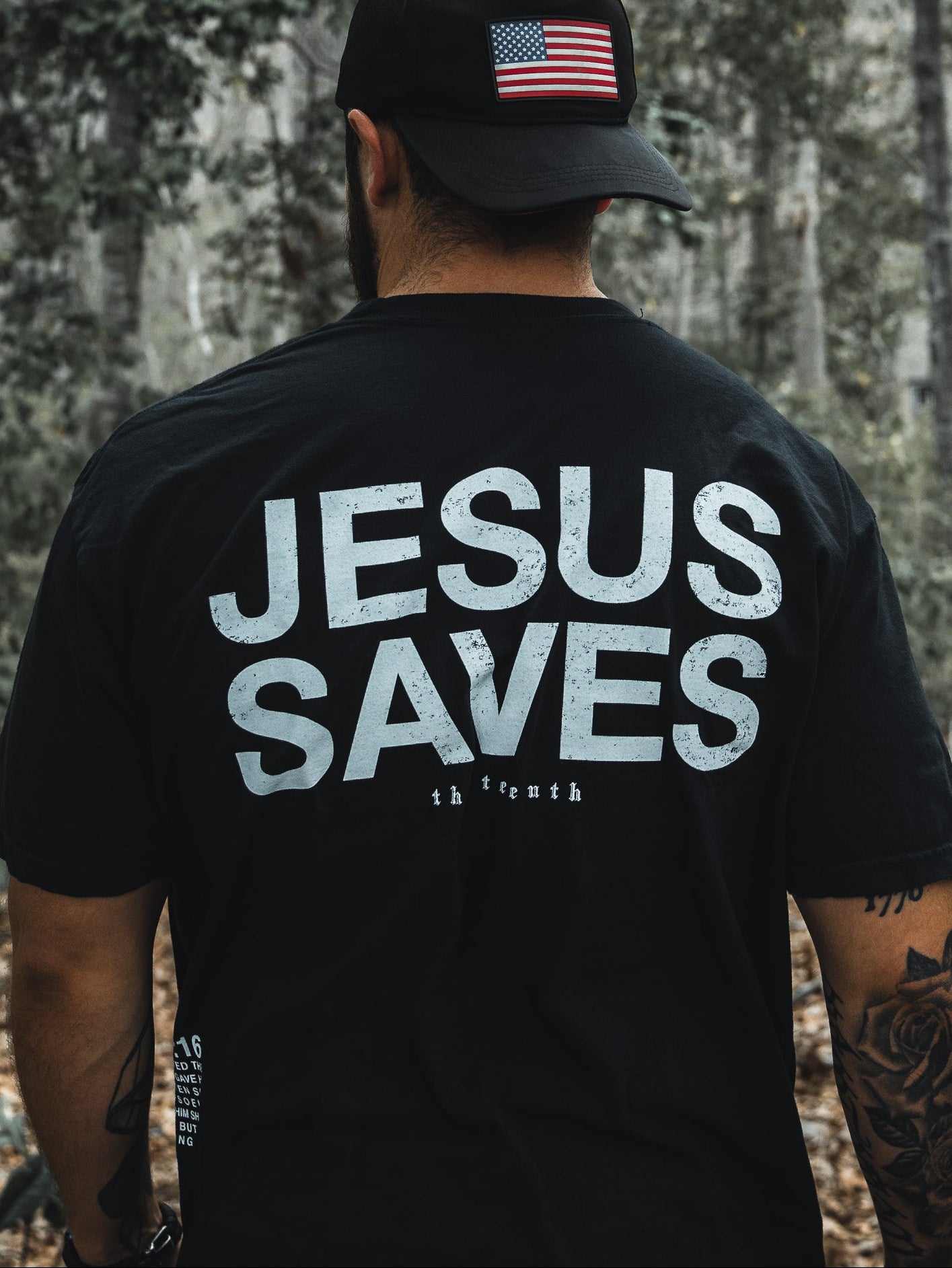 In a wooded area, a man dons the Jesus Saves - Black Tee by 13th and a black cap with an American flag. His tattooed arms suggest his deep faith, possibly inspired by John 3:16s promise of everlasting life through the begotten son.