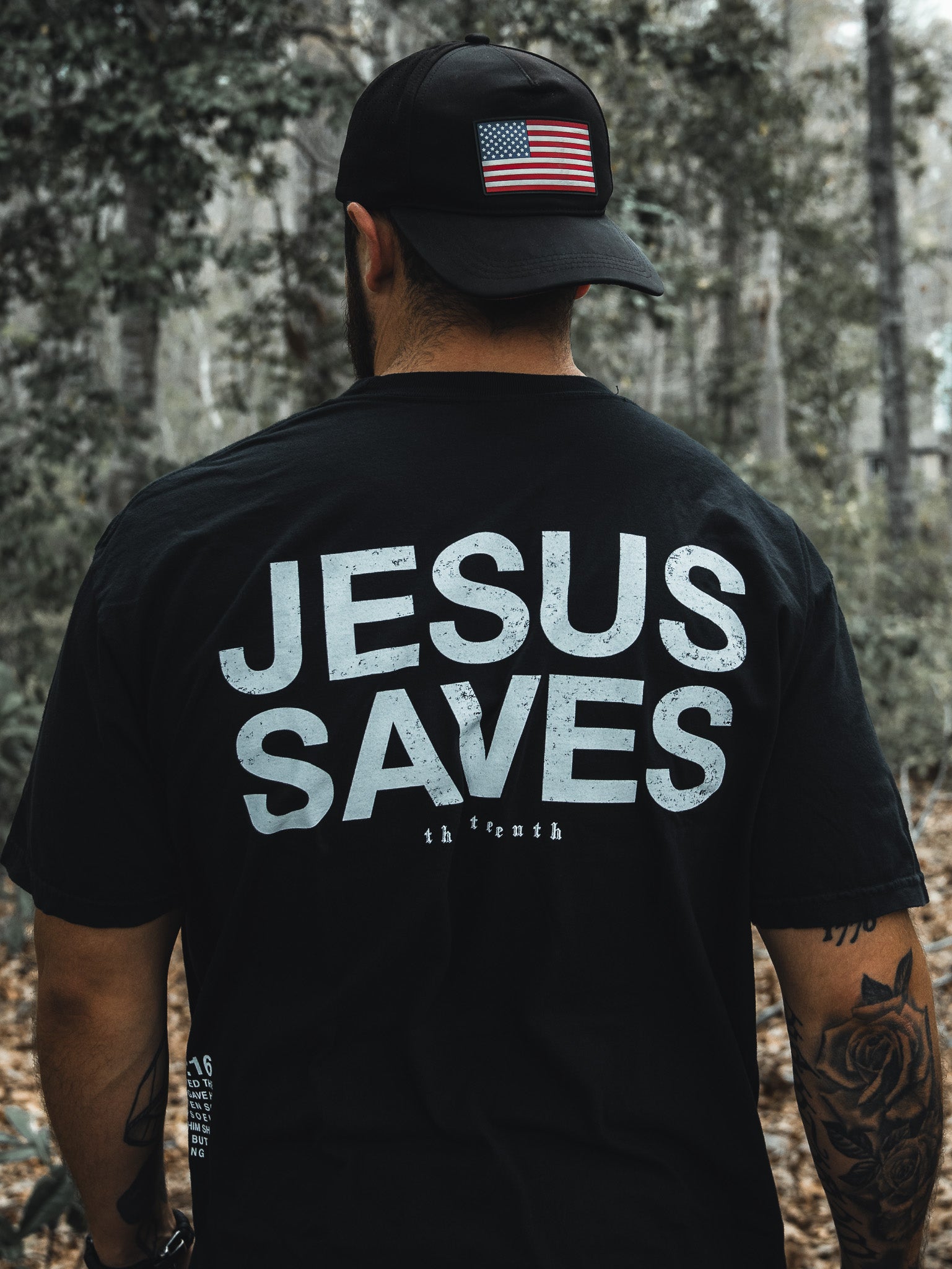 In a wooded area, a man dons the Jesus Saves - Black Tee by 13th and a black cap with an American flag. His tattooed arms suggest his deep faith, possibly inspired by John 3:16s promise of everlasting life through the begotten son.