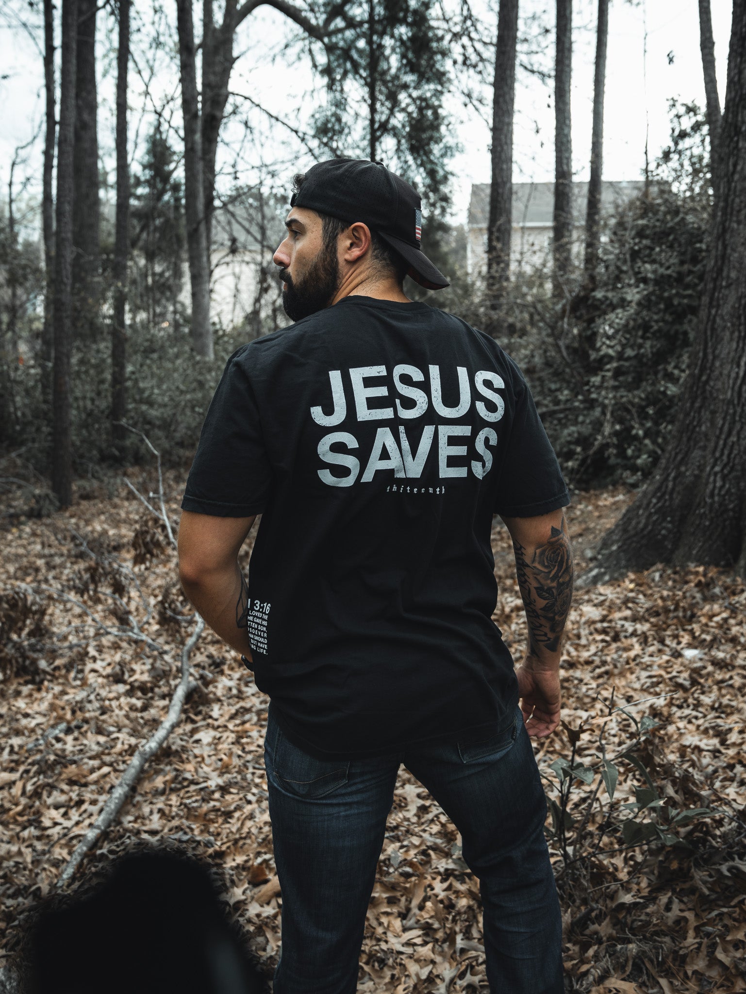 In a forest setting, a bearded man dons the 13th brands Jesus Saves - Black Tee featuring JESUS SAVES on the back. His sideways gaze amidst towering trees and leafy ground reflects the John 3:16-inspired message of eternal life.