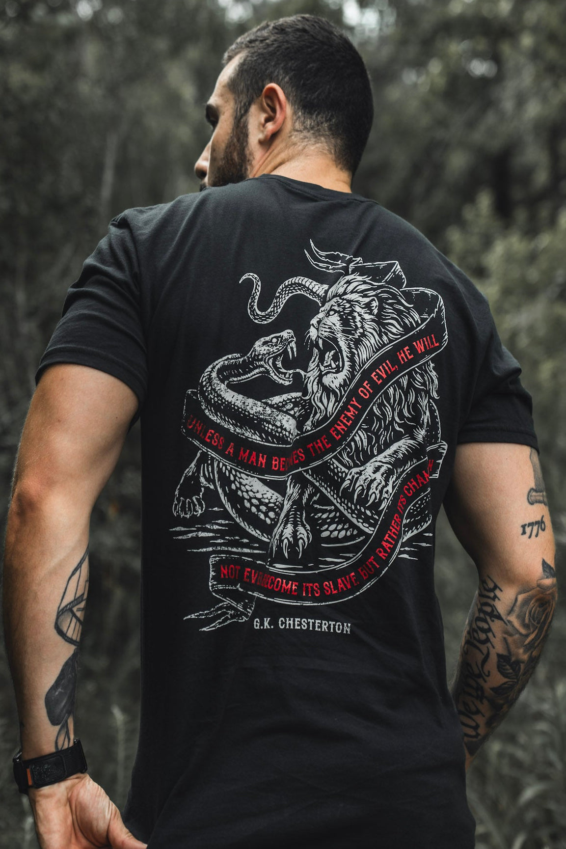 A tattooed man stands outdoors, facing away, wearing The Last Stand tee by Born United Private Label—black with a lion and snake graphic, red circular border, and G.K. Chesterton quote. Blurred trees fill the background.
