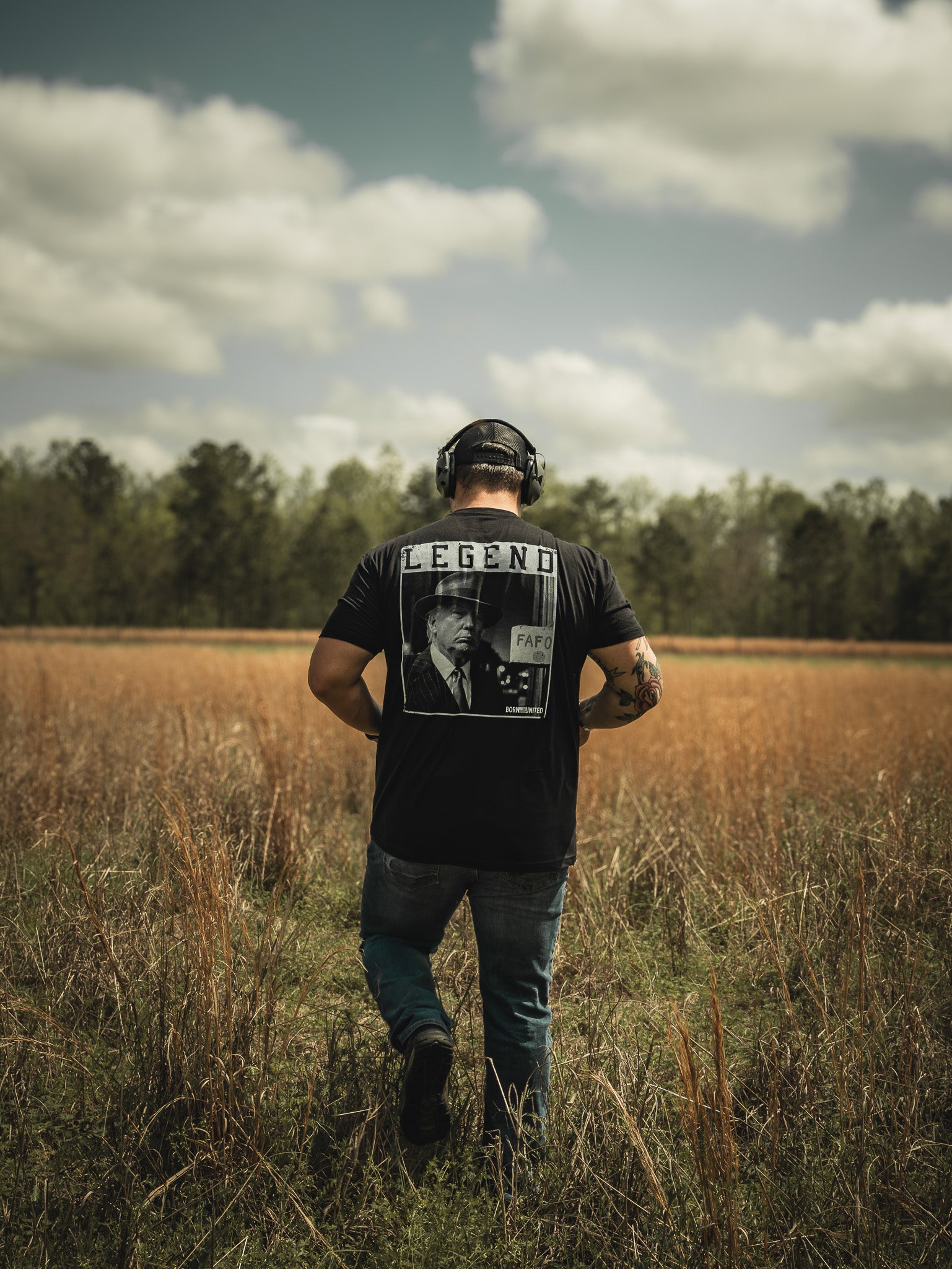 A man with headphones, sporting Born United Private Labels Legend FAFO black graphic T-shirt, strolls through a grassy field under a partly cloudy sky with trees in the background.