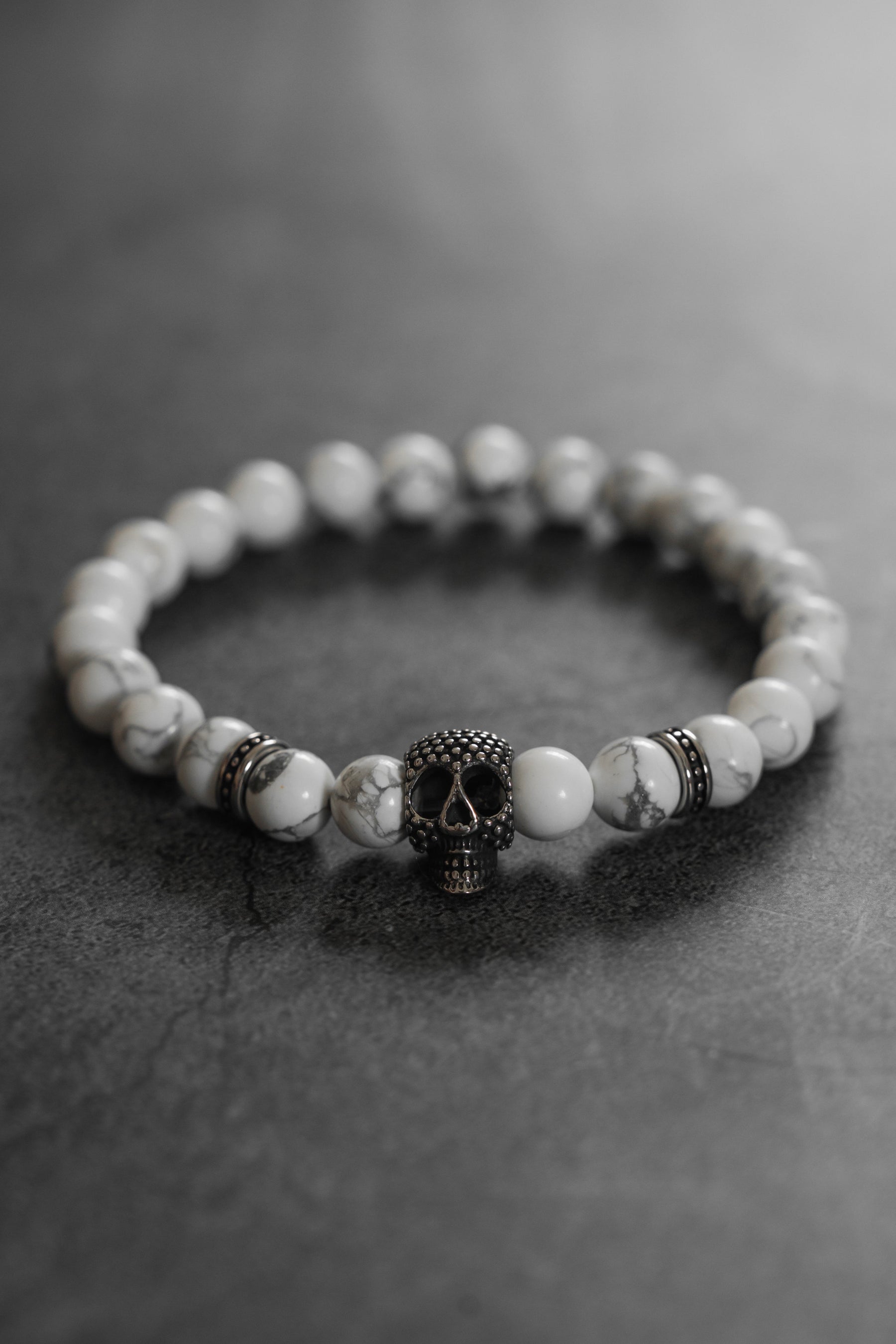 The Until Death Storm Cloud Bead Bracelet by Relentless Betrayal showcases white marbled beads and a bold metallic skull centerpiece, stylishly presented on a dark, textured surface.