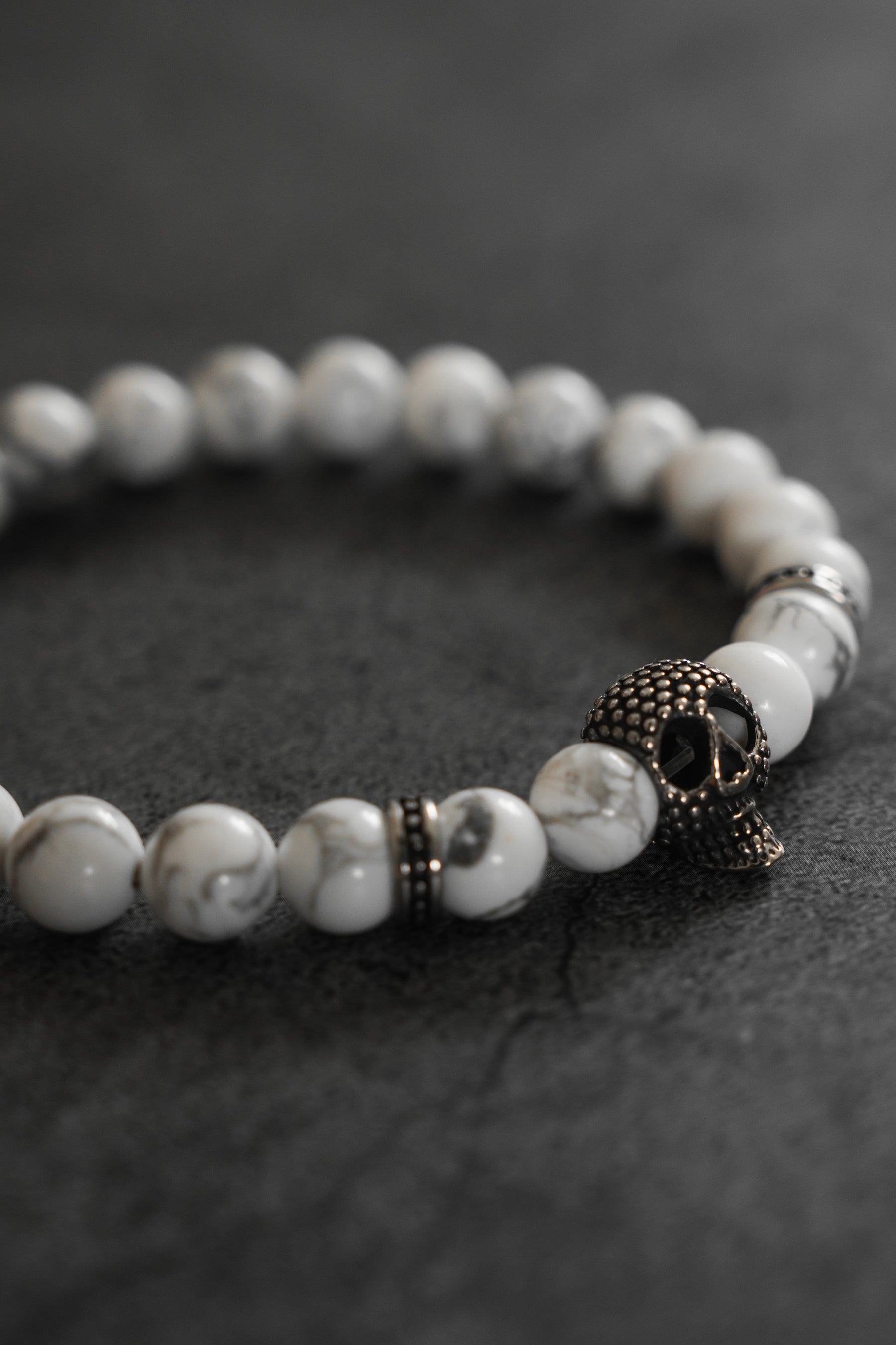 The Relentless Betrayal Until Death Storm Cloud Bead Bracelet, featuring white marbled beads and a metallic skull charm, is displayed on a dark, textured surface.