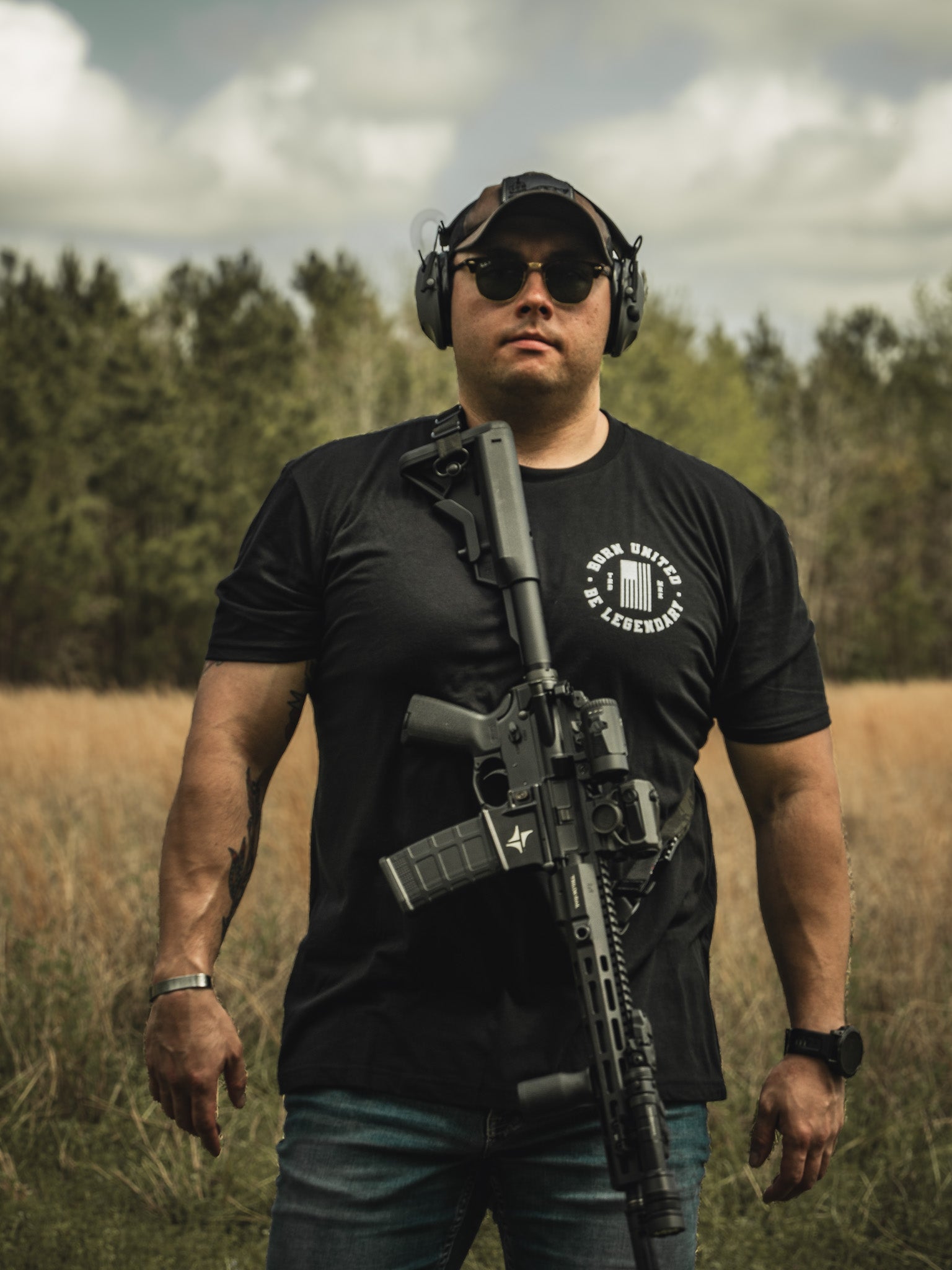 In a grassy outdoor area with trees and clouds, a person stands holding a Legend FAFO rifle by Born United Private Label, wearing sunglasses, headphones, and a black T-shirt.