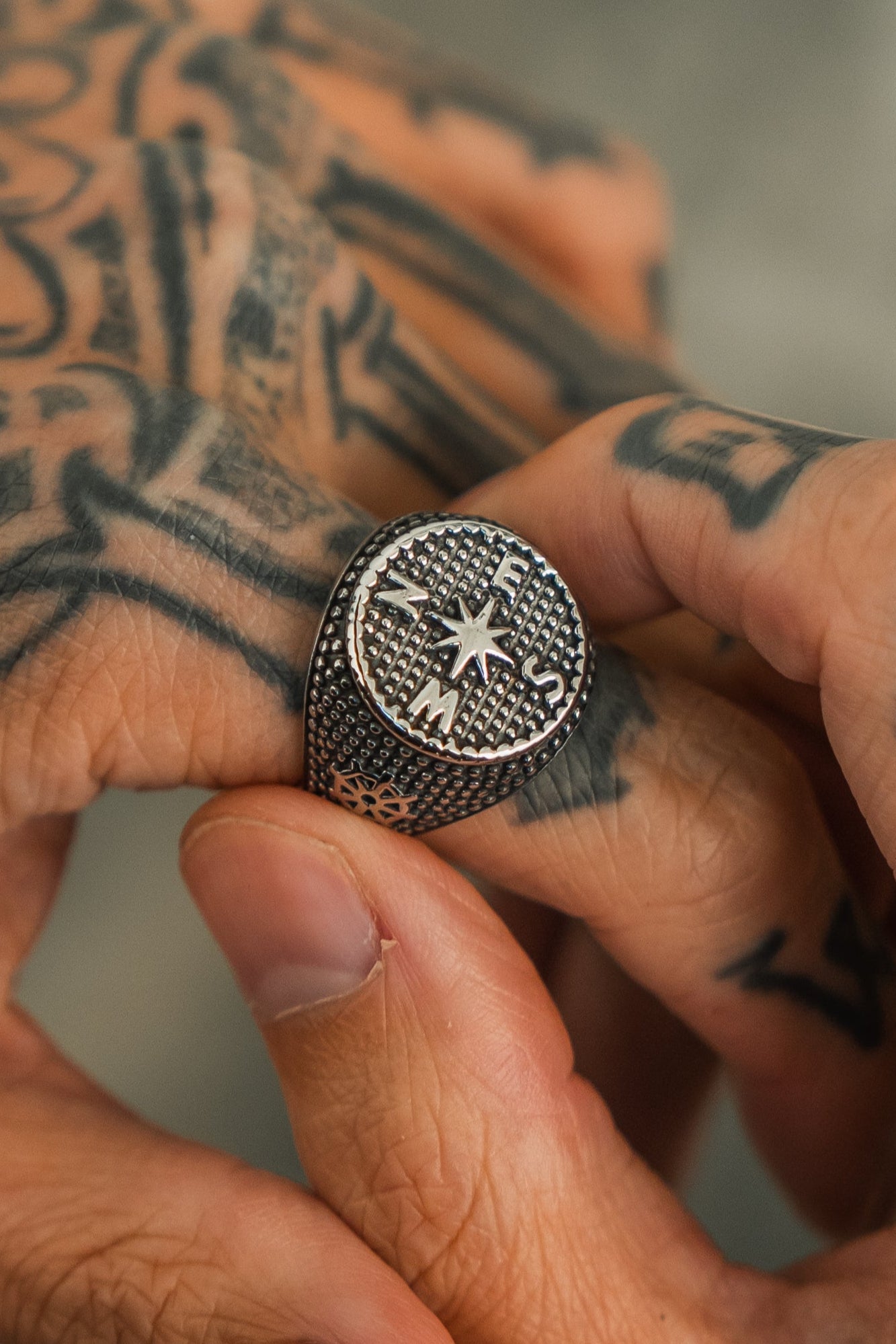 Someone with tattooed hands wears the handcrafted Crossroads Ring from Relentless Betrayal. Its textured stainless steel design features a compass with E, S, and M directions, complementing the intricate tattoos on their hand beautifully.