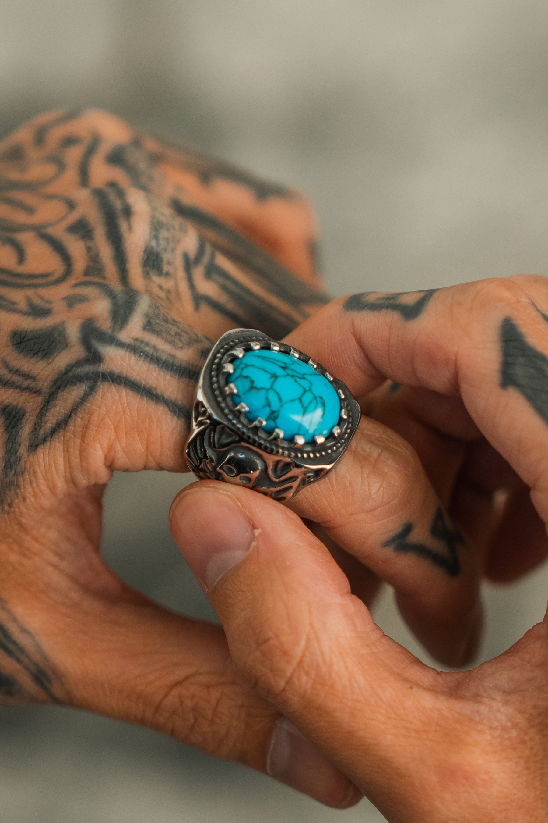 A tattooed hand displays the Outsider Ring by Relentless Betrayal, featuring a vibrant turquoise stone and intricate metalwork, set against a softly blurred background.