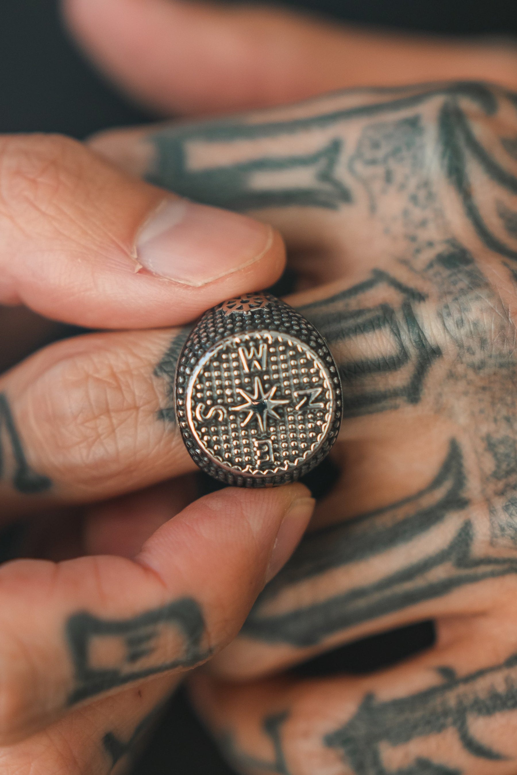 A persons tattooed hand holds the Crossroads Ring by Relentless Betrayal, an exquisite stainless steel piece featuring a compass design with N, E, S, and W directions. A textured dotted pattern enhances its unique allure.