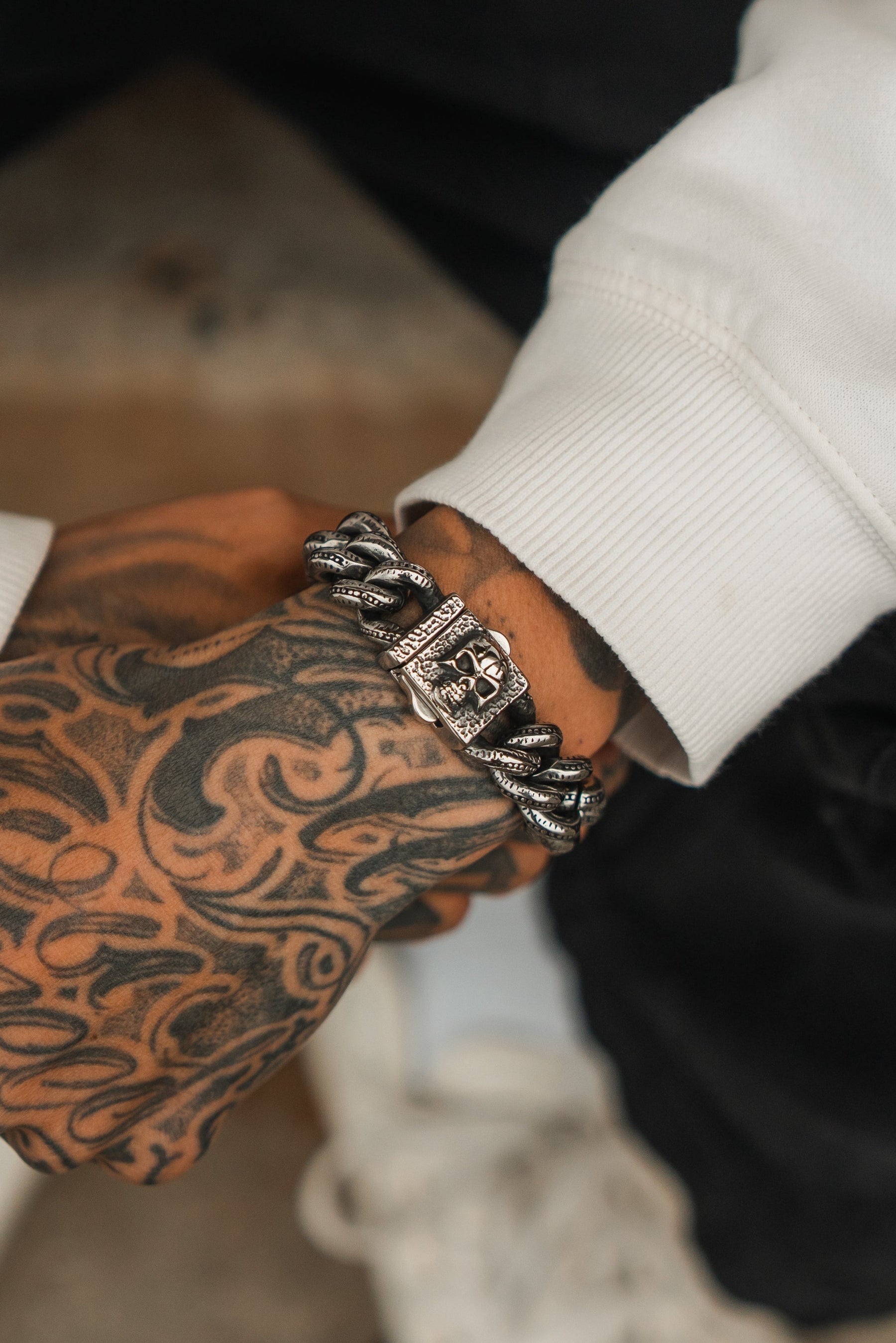 A close-up shows a tattooed forearm and hand wearing Relentless Betrayals chunky Tombstone Bracelet. Dressed in a long-sleeved white shirt, the focus highlights both the intricate tattoos and the distinctive bracelet design.
