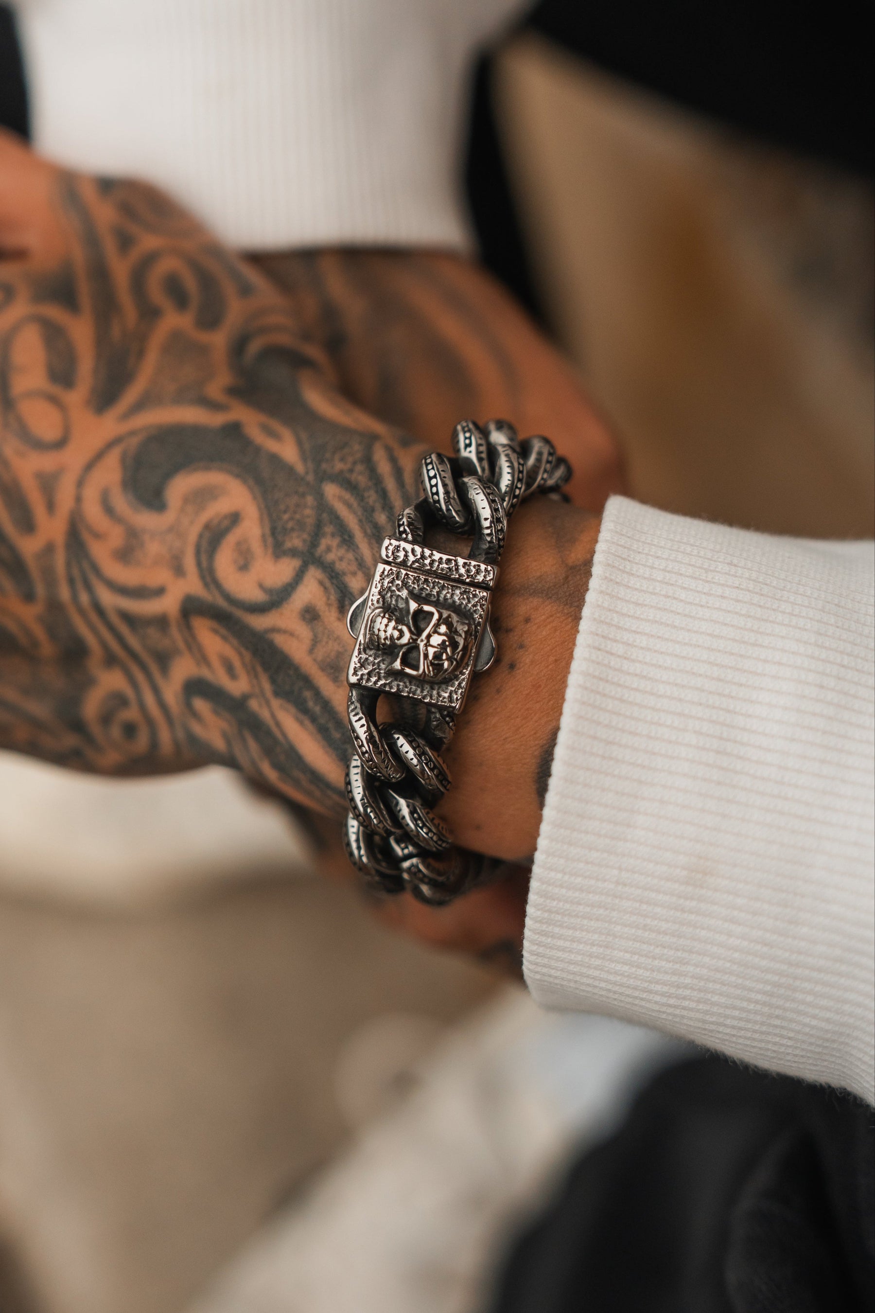 A close-up of a tattooed hand wearing a chunky Relentless Betrayals Tombstone Bracelet crafted from stainless steel, featuring intricate designs and a square clasp. The bracelets decorative patterns rest elegantly against a white sleeve.