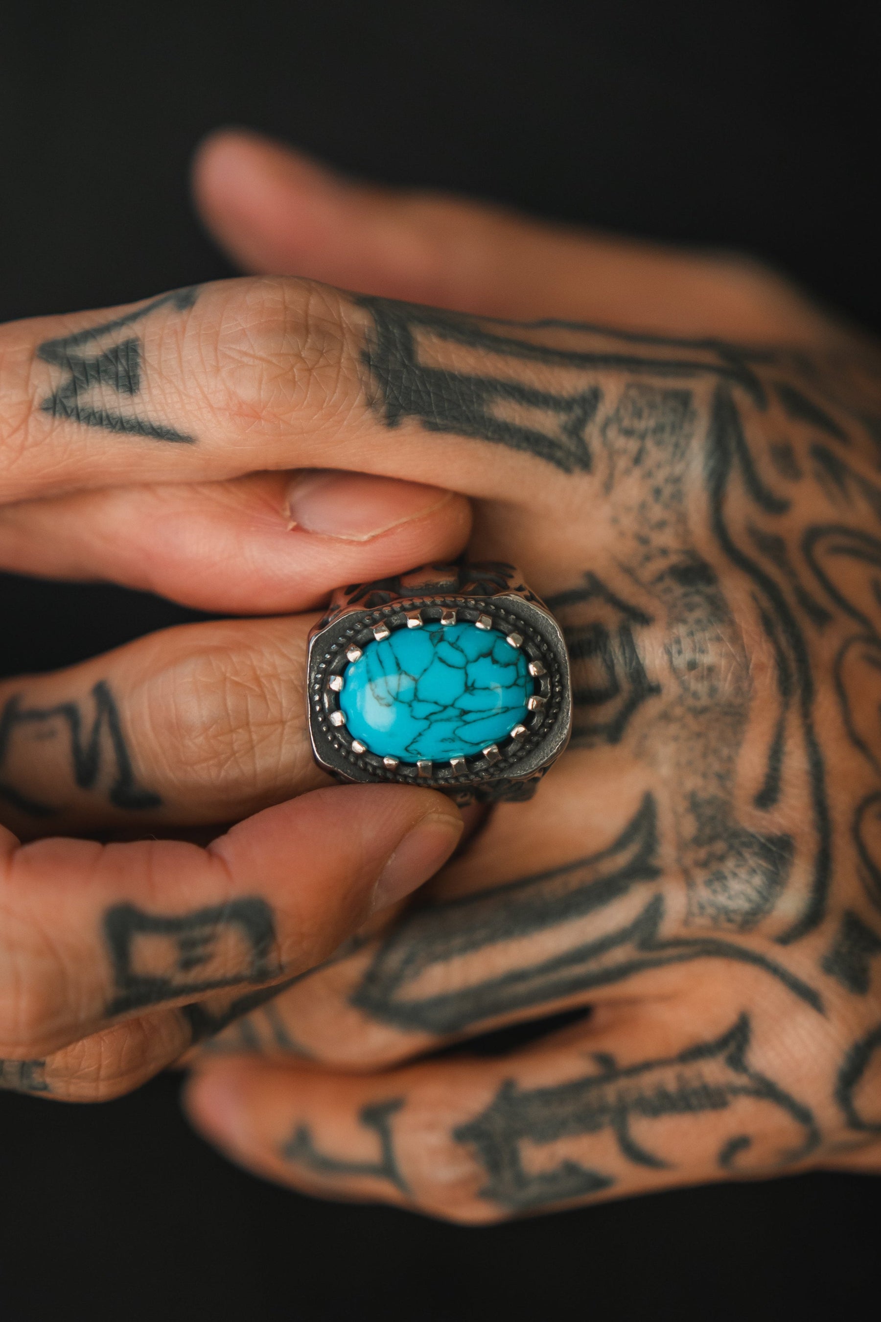 A tattooed hand showcases Relentless Betrayals Outsider Ring, featuring a handcrafted silver band with a large oval turquoise stone accented by black veins. The intricate skin art enhances the detailed metalwork and timeless allure of this unique ring.