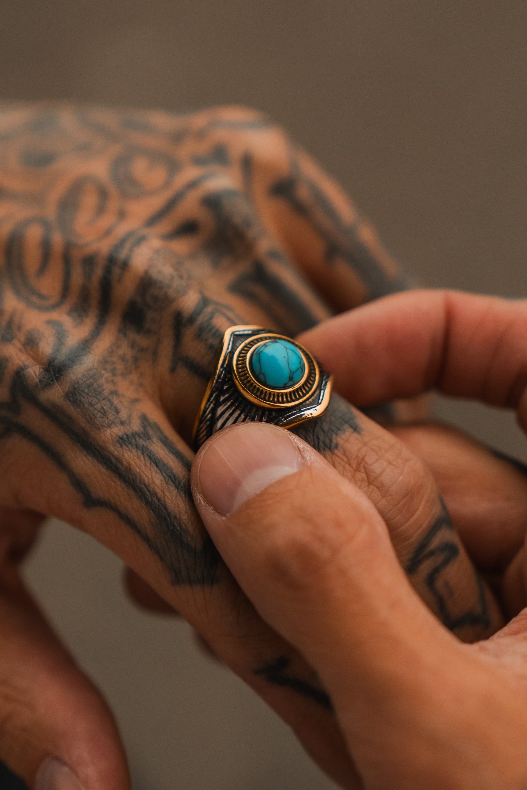 A tattooed hand wears Relentless Betrayals handcrafted Royal Crest Ring featuring a turquoise stone, gold accents, and stainless steel. Another hand gently holds the tattooed one, highlighting the unique jewelry and tattoos.