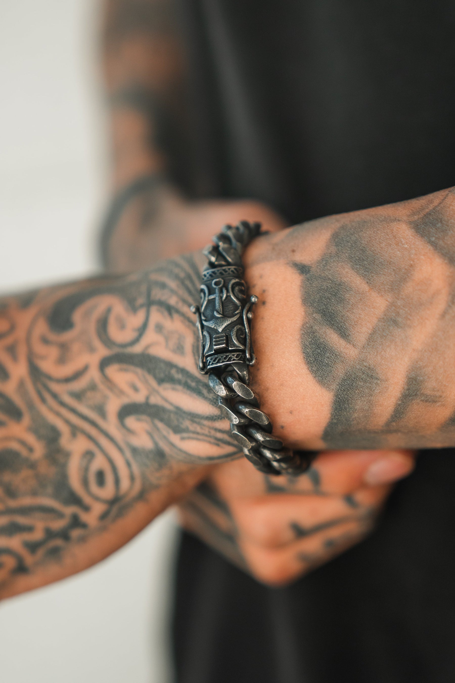 A close-up shows a person wearing Relentless Betrayals handcrafted Voyager Bracelet, featuring a carved pattern. Their tattooed arm displays intricate maritime designs, paired with a dark shirt.