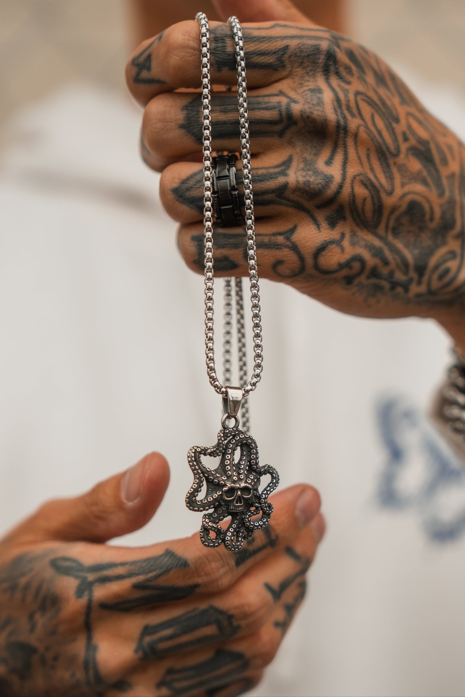 A tattooed individual in a white hoodie holds the Depths of the Sea Necklace by Relentless Betrayal, featuring a silver chain with an octopus-skull pendant inspired by bioluminescent sea creatures.