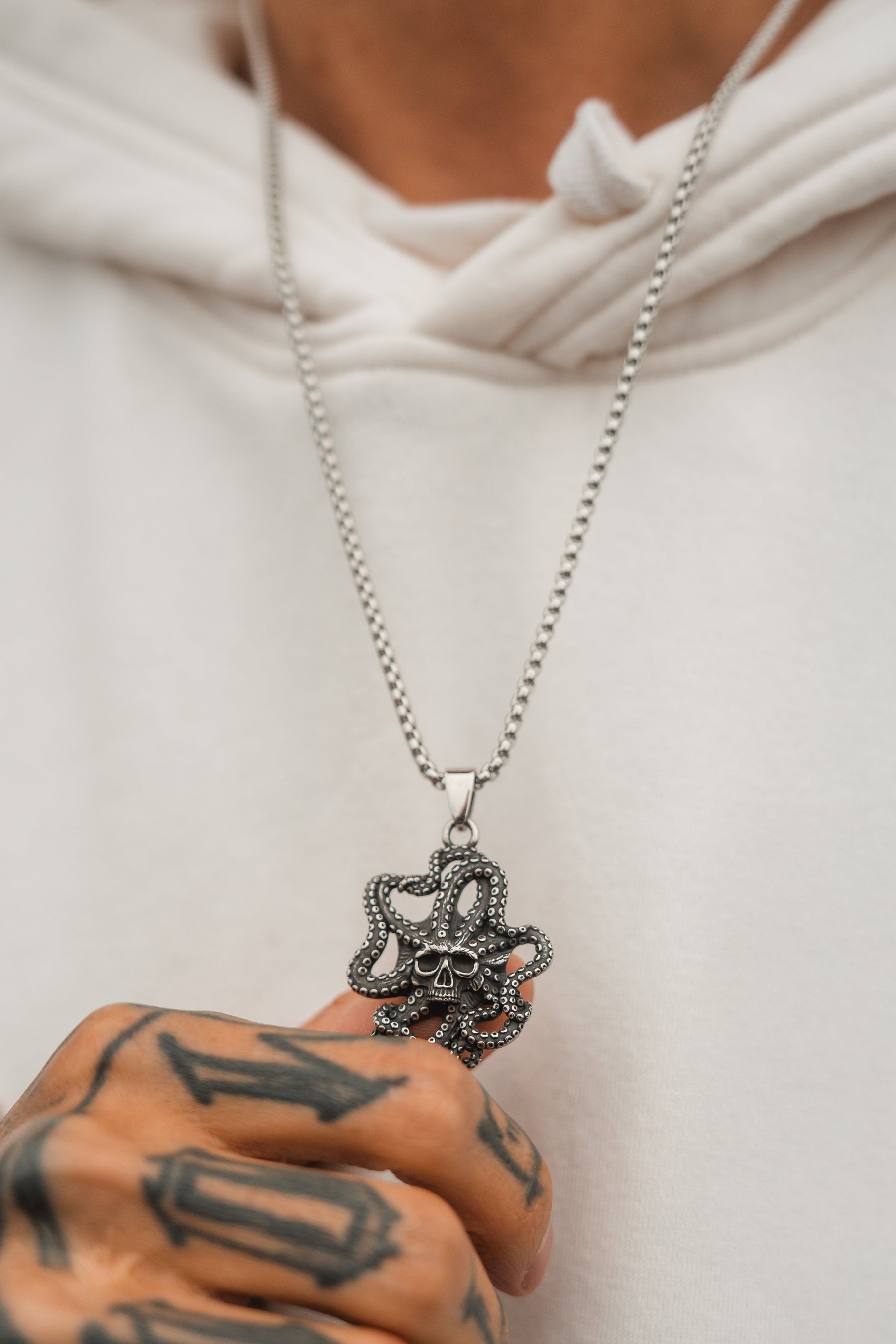 A person in a white hoodie grips the stainless steel chain of the Depths of the Sea Necklace by Relentless Betrayal, with their tattooed hand showcasing geometric designs and numbers. The softly blurred background adds an air of mystery.