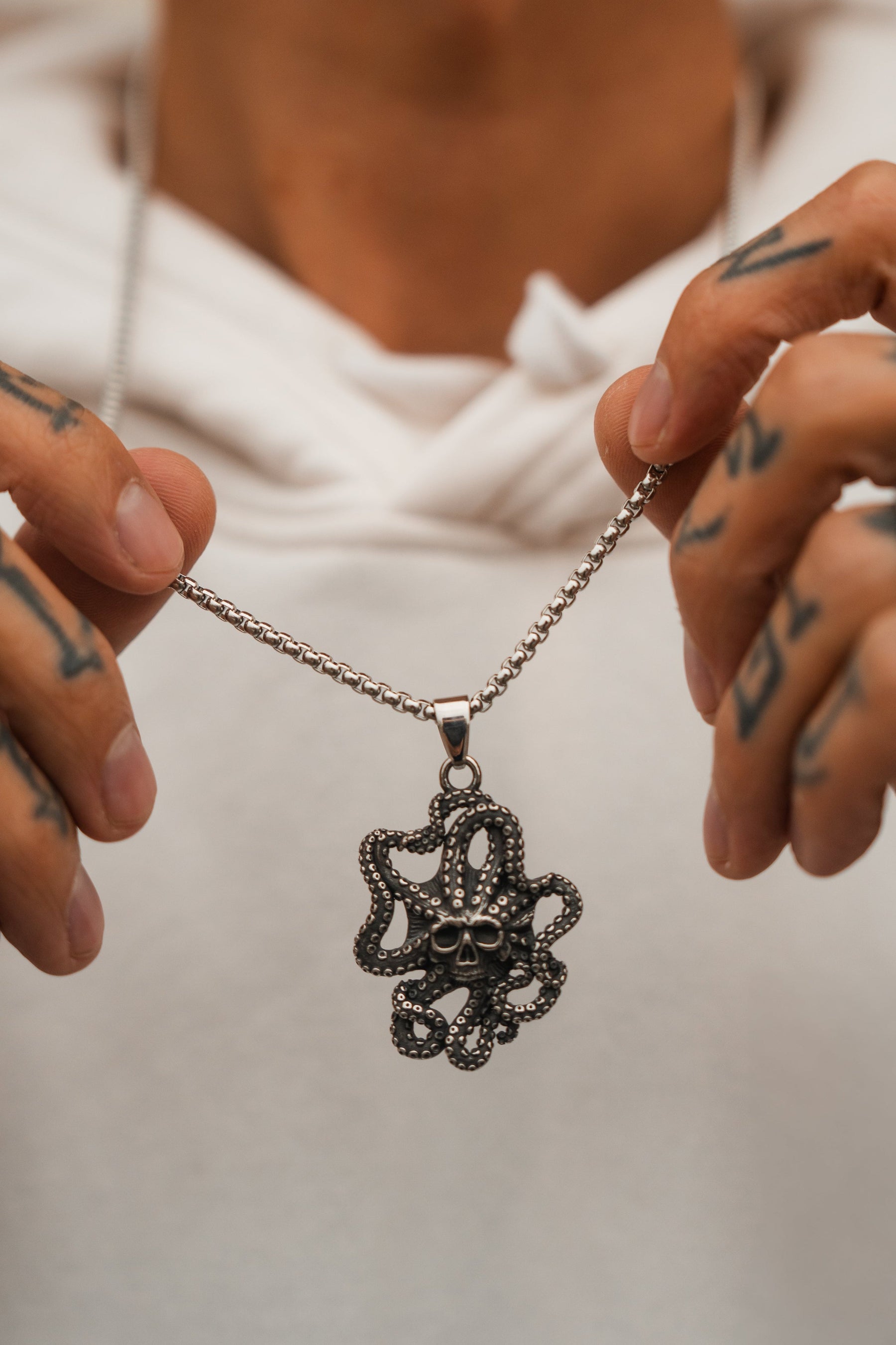 A tattooed person in a white hoodie holds the Relentless Betrayals Depths of the Sea Necklace, detailed with a stainless steel chain and an octopus pendant, featuring intricate tentacles around a small skull, reminiscent of oceanic bioluminescent creatures.