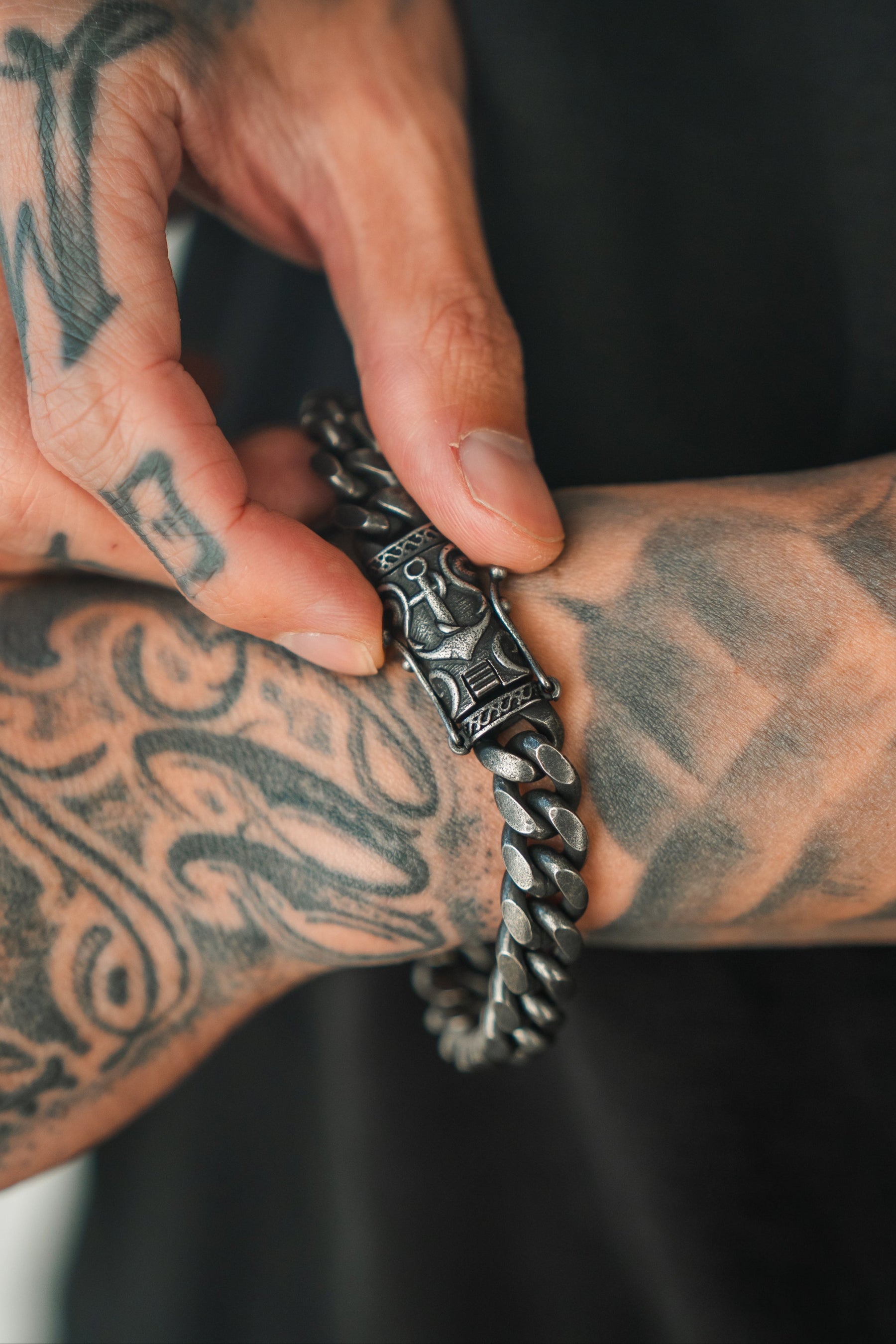 A tattooed hand adjusts the Voyager Bracelet by Relentless Betrayal, showcasing exquisite handcrafted jewelry with abstract and tribal designs. This chunky metal piece features an engraved clasp—ideal for maritime journeys.