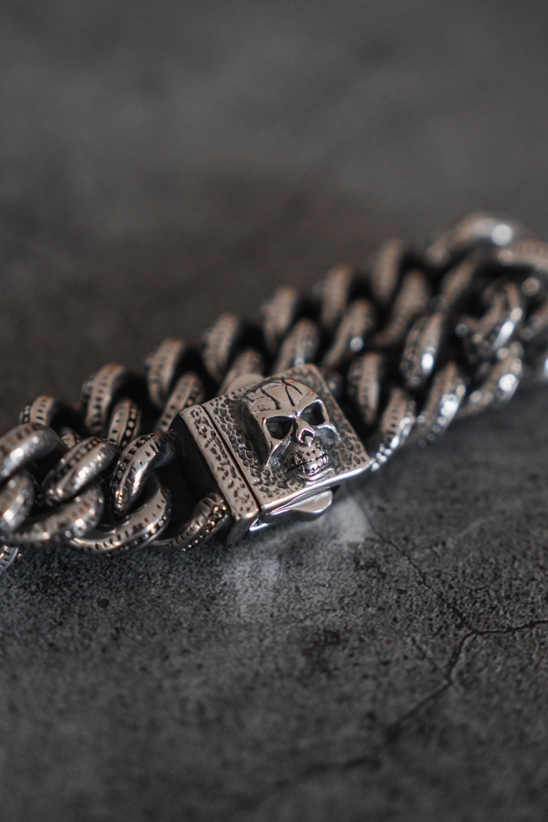 A close-up of Relentless Betrayals The Tombstone Bracelet showcases a textured metal masterpiece with a skull clasp on a dark background. Its large, intricately detailed links in polished stainless steel exude a rugged, gothic aesthetic.