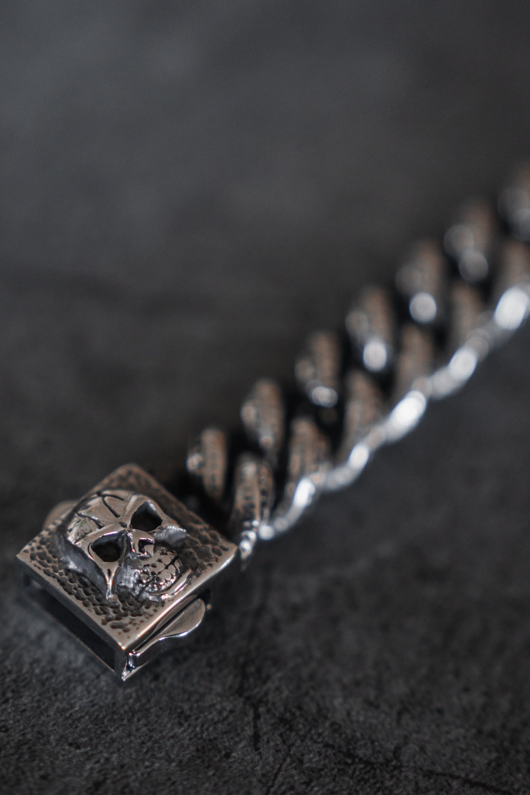 Close-up of The Tombstone Bracelet by Relentless Betrayal, showcasing a stainless steel metallic chain featuring a detailed skull on one square link. Its texture and design stand out against a dark background, evoking remembrance.