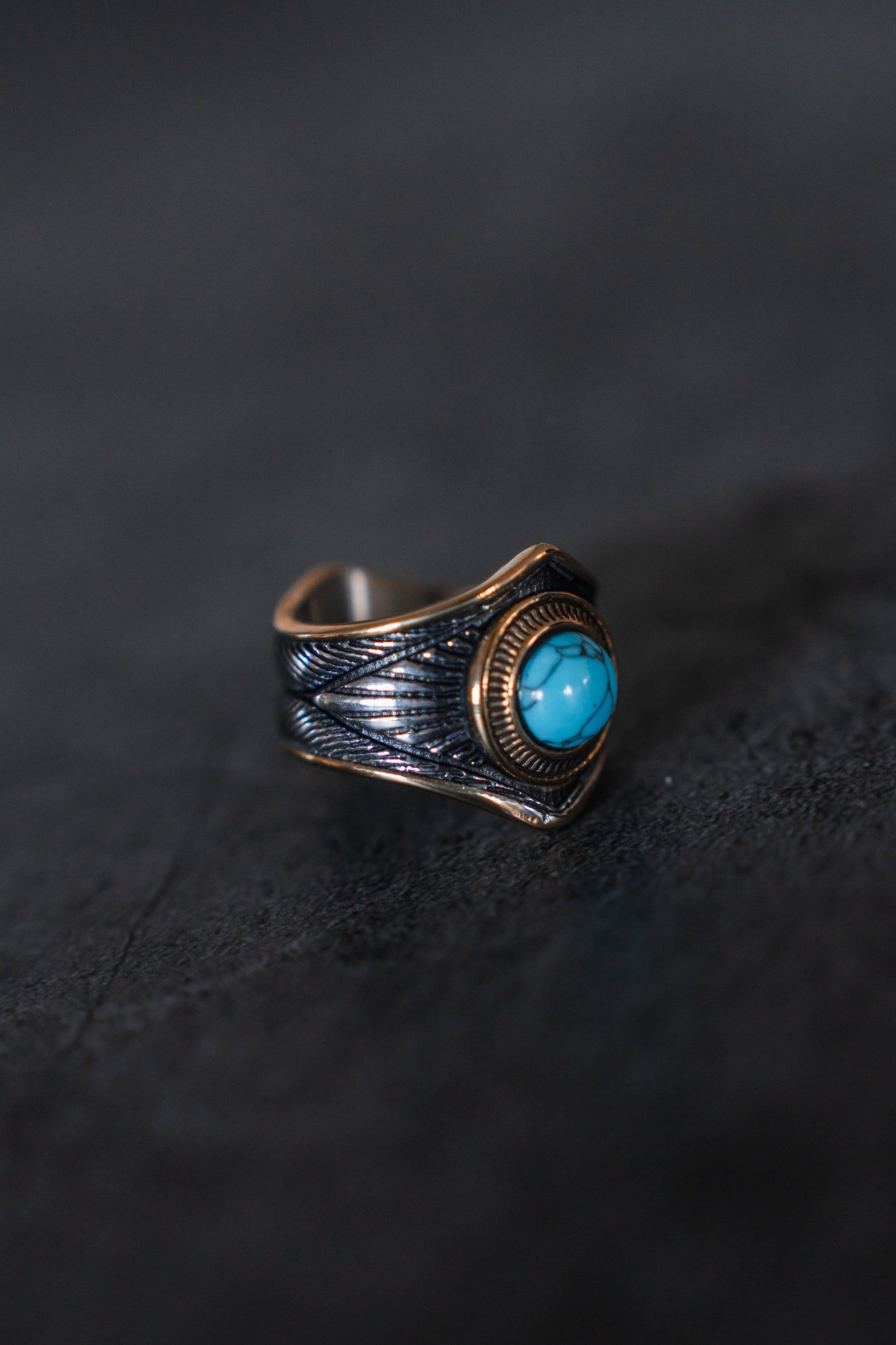 The handcrafted The Royal Crest Ring by Relentless Betrayal features intricate stainless steel patterns and a central turquoise stone, elegantly displayed on a dark textured surface.