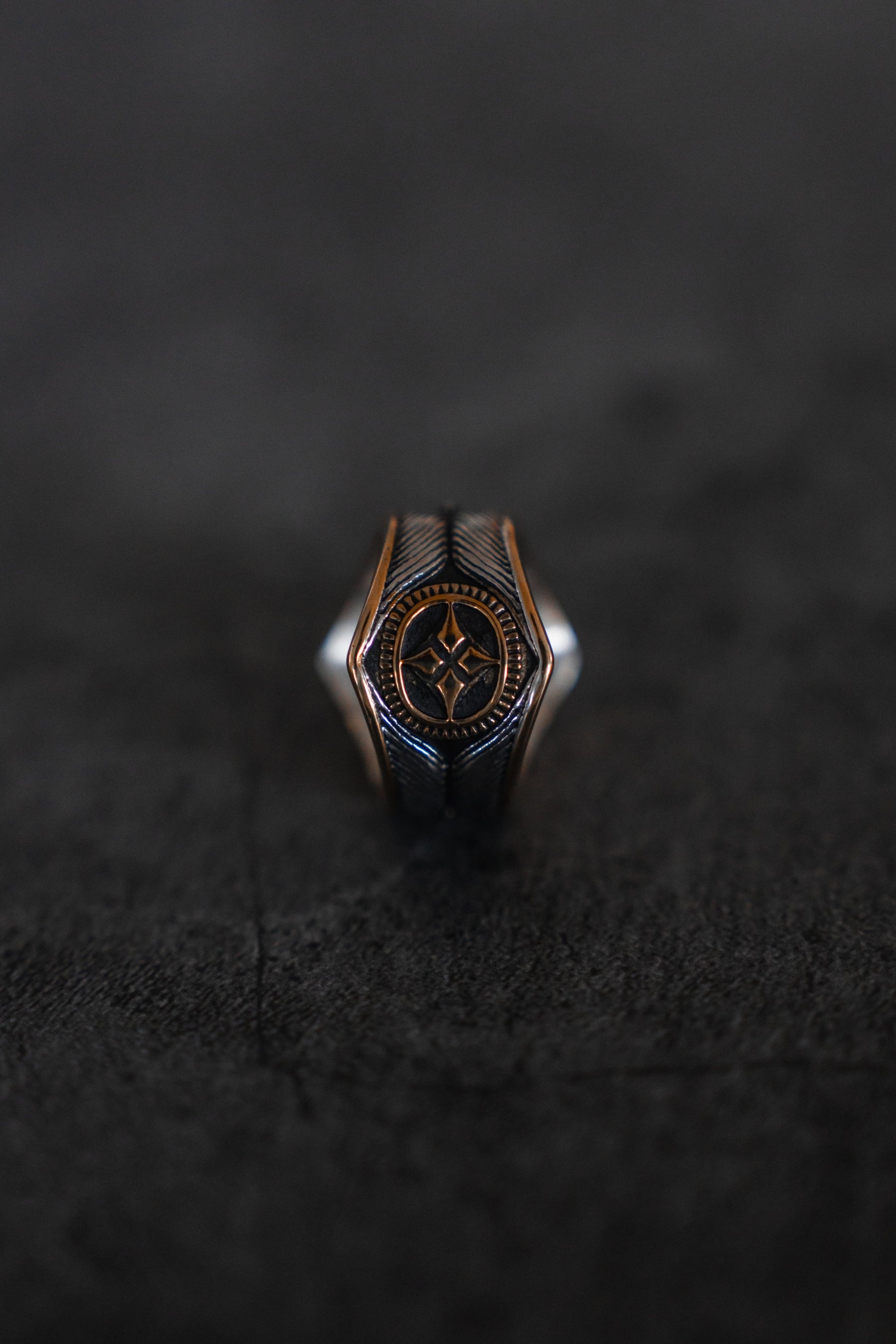 The Royal Crest Ring by Relentless Betrayal showcases an intricate star-like design on a dark textured surface. Handcrafted with silver and bronze, its blend of metallic shades emphasizes exquisite craftsmanship, giving it antique charm and timeless appeal.