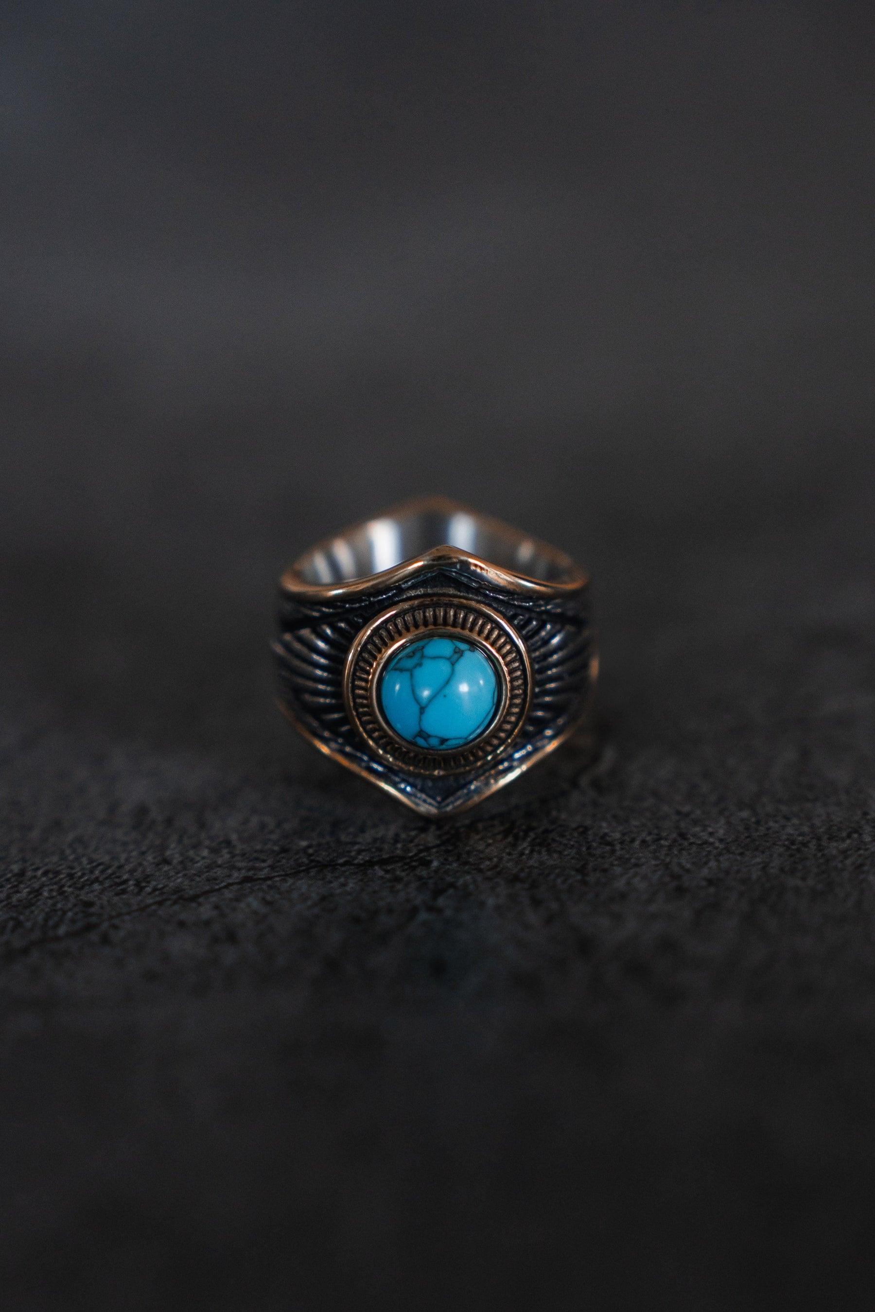 The Royal Crest Ring by Relentless Betrayal is a handcrafted silver and bronze piece with detailed designs. It boasts a round turquoise stone in its center, set against an intricately dark textured background.