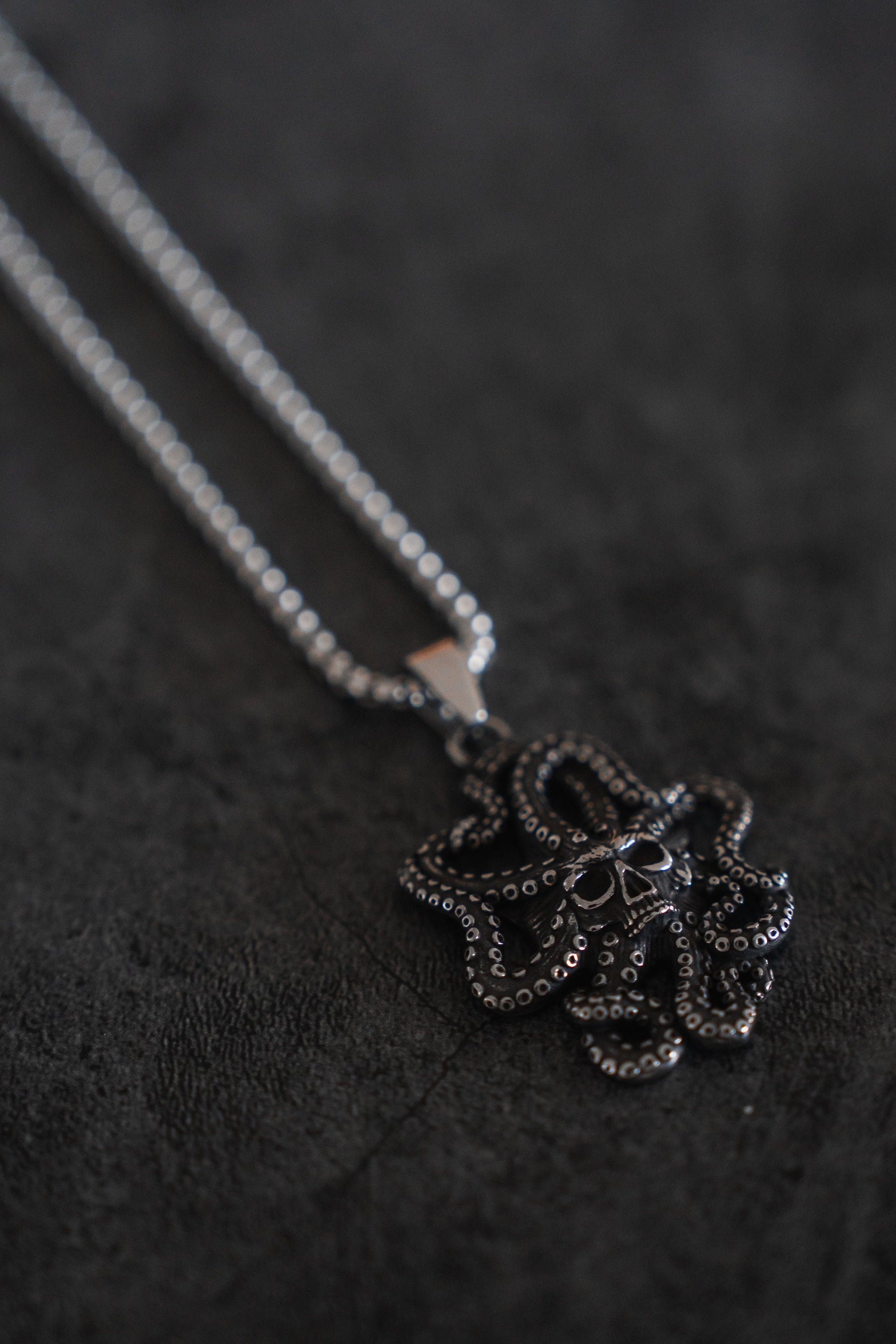 The Depths of the Sea Necklace by Relentless Betrayal, featuring a silver chain and an intricately detailed octopus pendant, lies on a dark textured surface, evoking the oceans depths with its striking contrast.