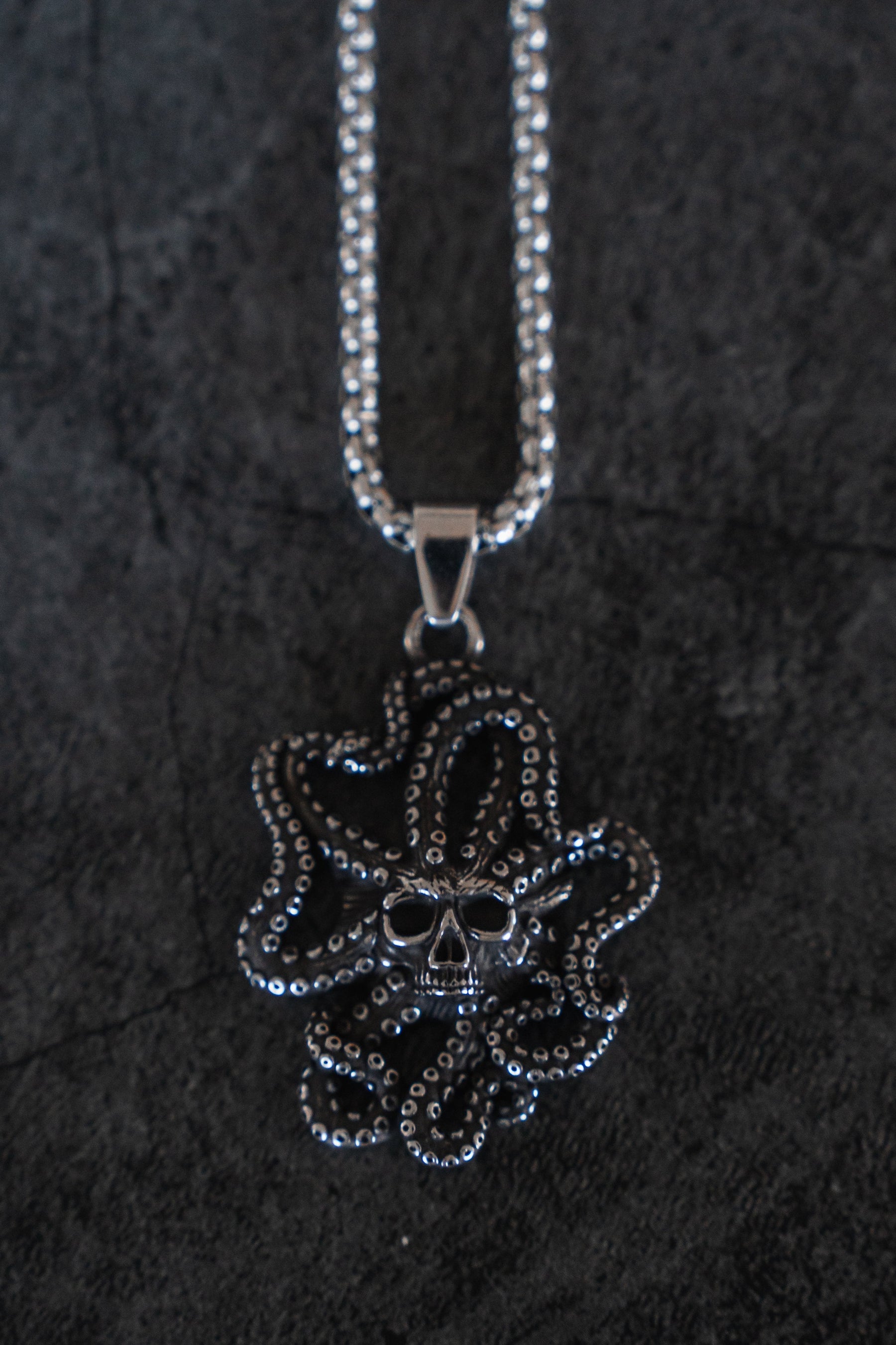 The Depths of the Sea Necklace by Relentless Betrayal is a silver stainless steel piece with a skull pendant entwined by an octopuss detailed tentacles, capturing the oceans mysterious allure against a dark, textured background.