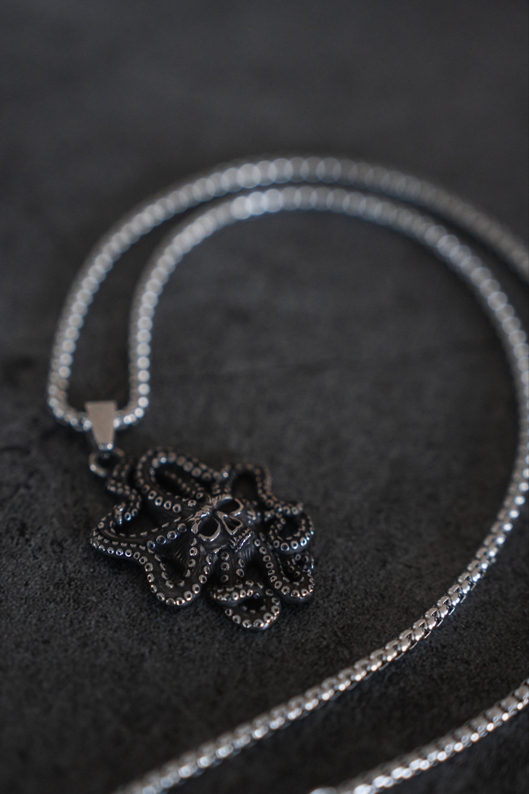 Relentless Betrayals Depths of the Sea Necklace features a detailed octopus pendant on silver and stainless steel, elegantly coiling on a dark gray surface to capture the oceans allure with its intricate patterns and texture.