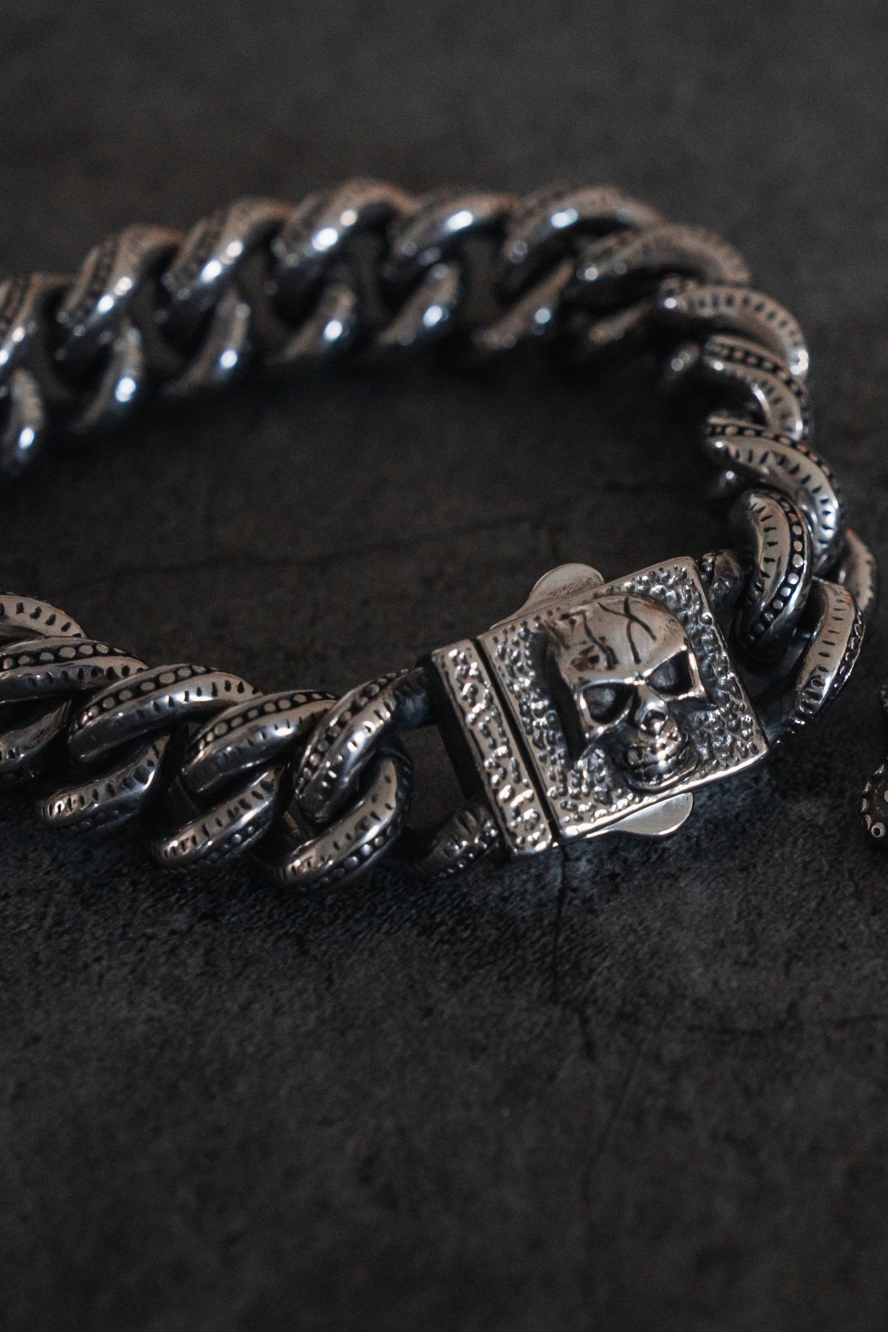 A close-up of The Tombstone Bracelet by Relentless Betrayal showcases a detailed skull design on its clasp. This intricately crafted stainless steel piece features textured links, elegantly resting on a dark surface.