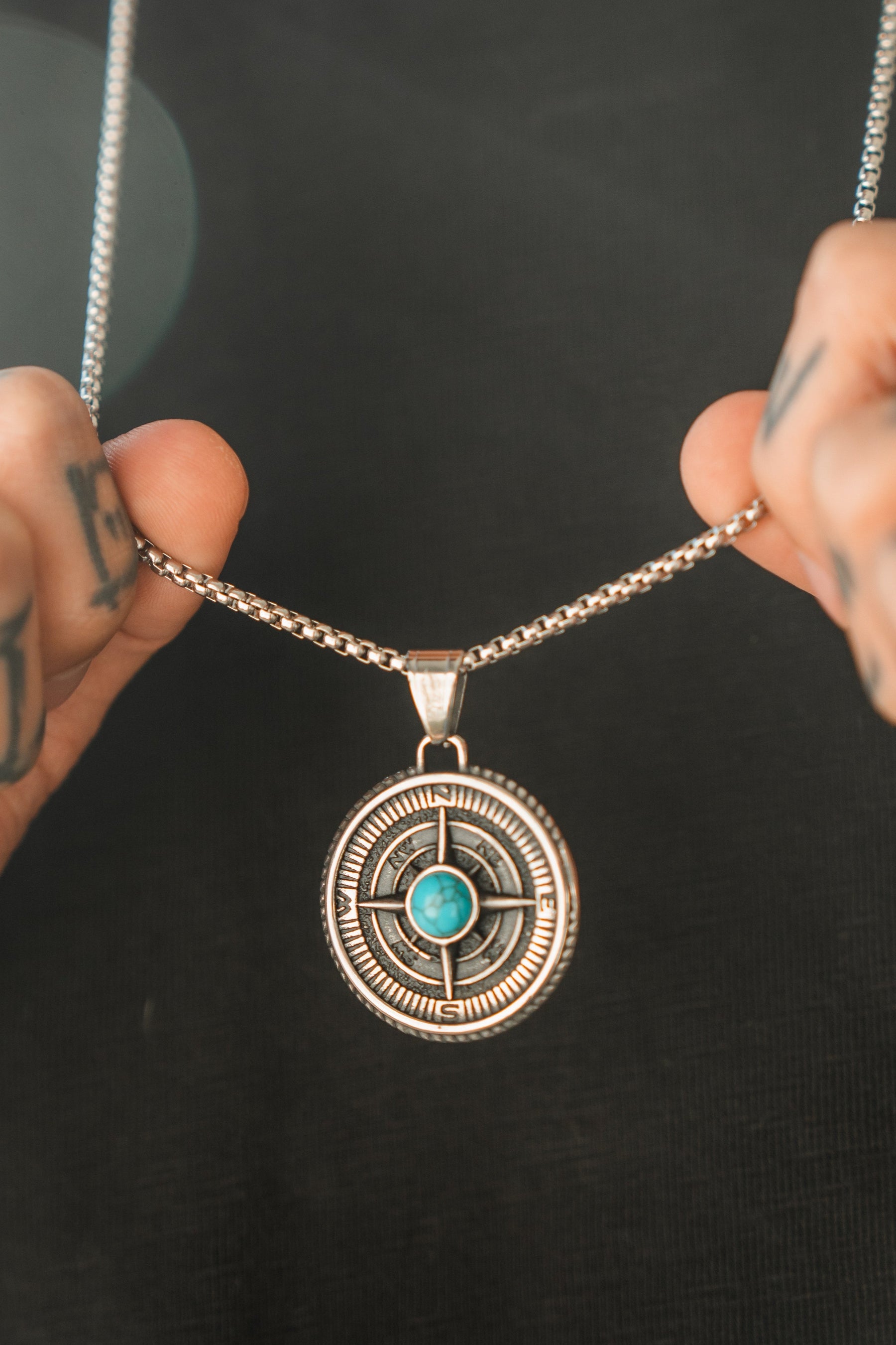 A person holds the Relentless Betrayal Crossroads Necklace, a silver chain with a round pendant featuring intricate designs and a turquoise stone, symbolizing transition. Their tattooed hands and dark shirt hint at pivotal moments in their journey.