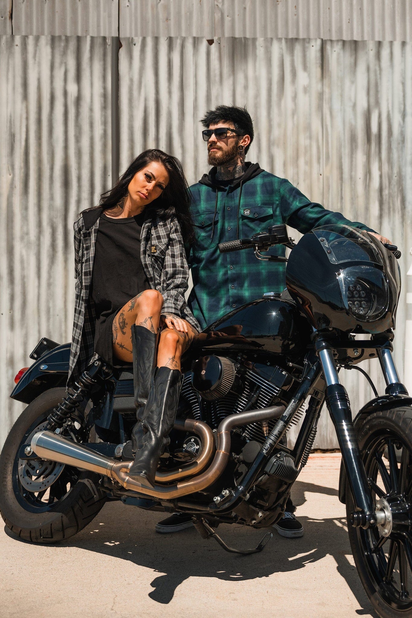 A woman in a black dress sits on a motorcycle as a man stands beside her wearing sunglasses and the Lost At Sea Premium Hooded Flannel by Relentless Betrayal. They exude an effortlessly cool vibe against a corrugated metal wall backdrop.