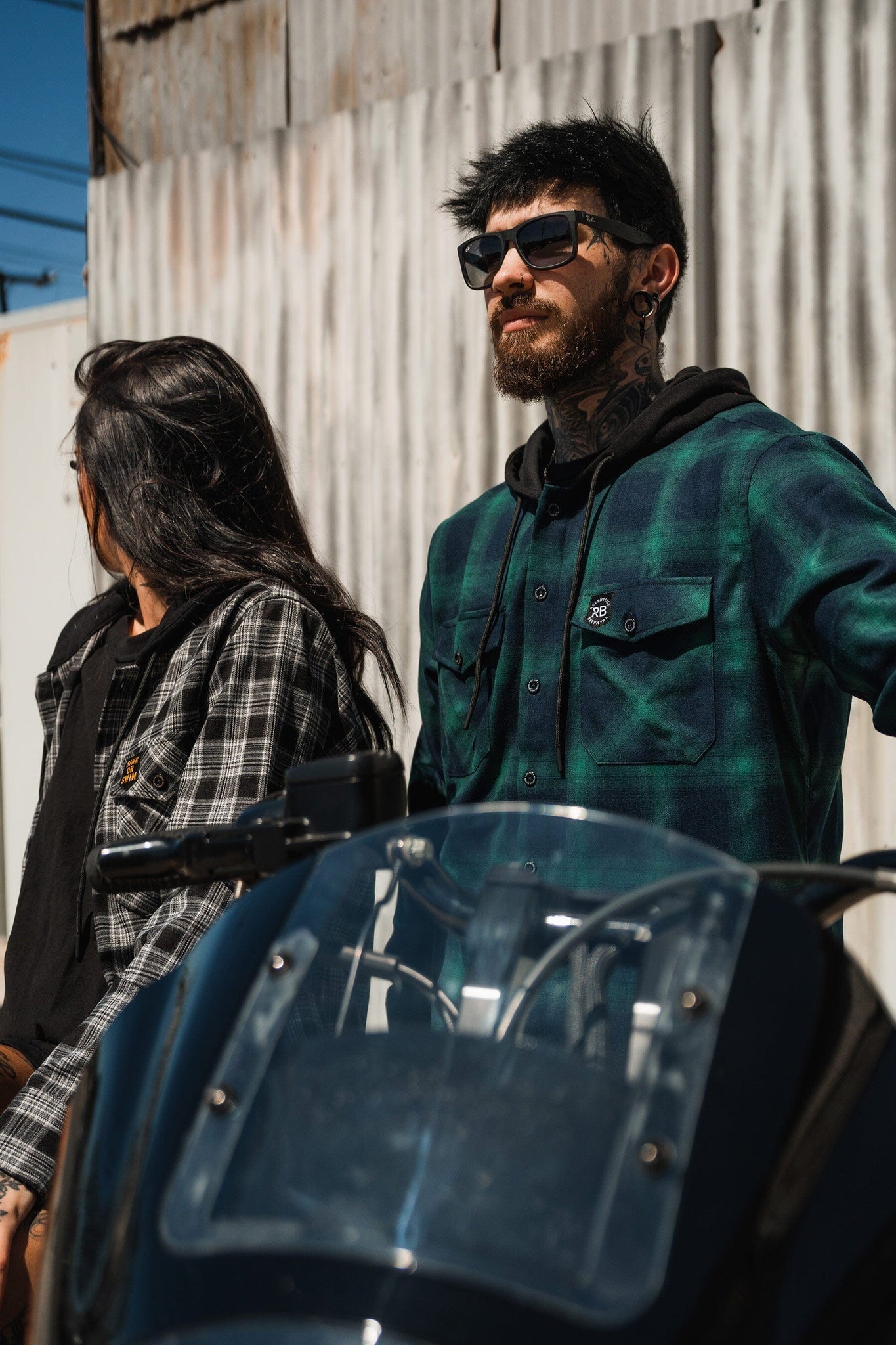 A tattooed man in sunglasses leans on a motorcycle, wearing a Lost At Sea Premium Hooded Flannel from Relentless Betrayal. Beside him, a woman with long dark hair dons another plaid shirt. They pose against a corrugated metal wall, showcasing the rugged charm of ultra-soft flannels.