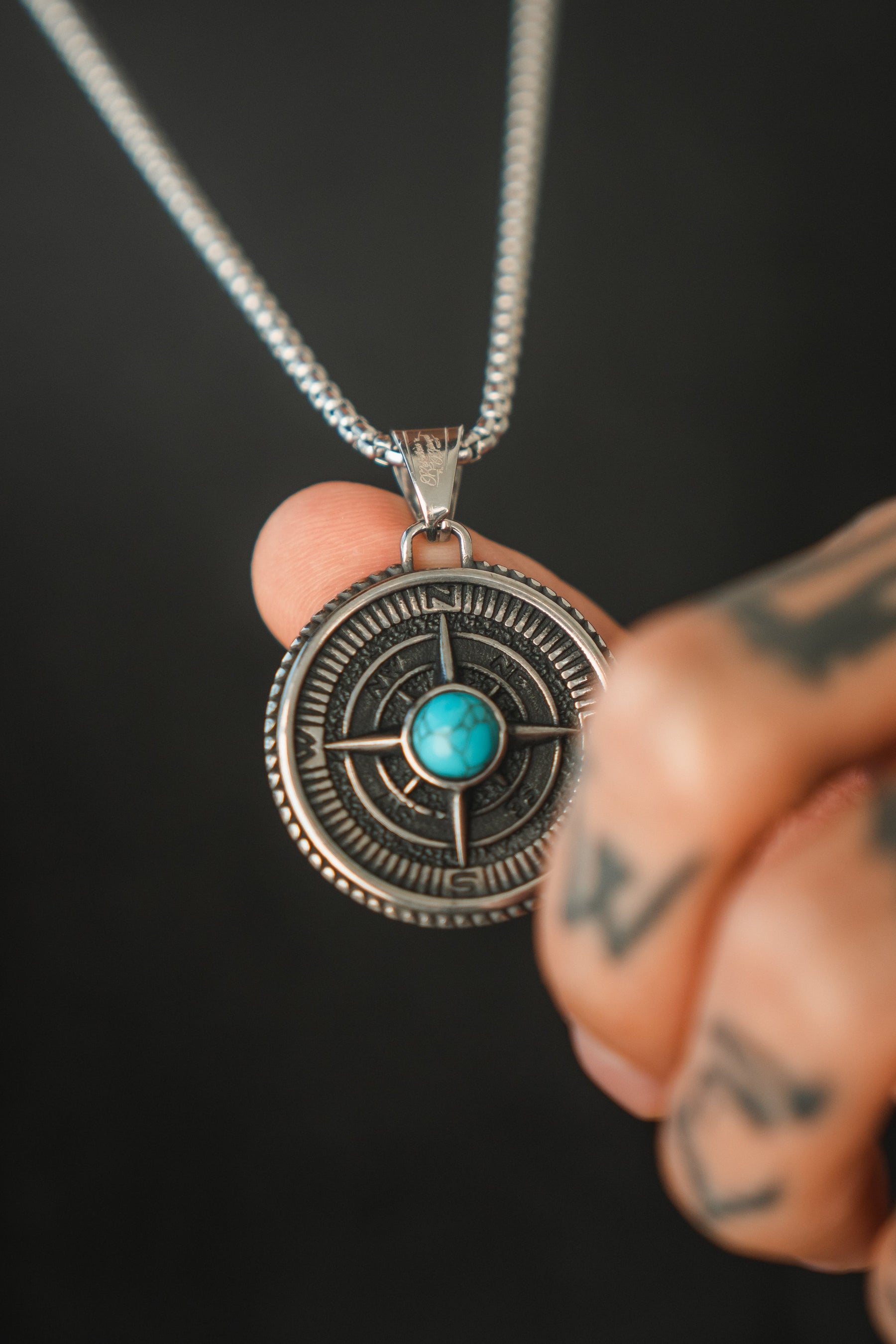 Tattooed fingers showcase the Crossroads Necklace by Relentless Betrayal, featuring a silver compass pendant with a turquoise center, symbolizing transition. Its intricate design and visible chain pop against a dark background, making it ideal for commemorating pivotal moments.