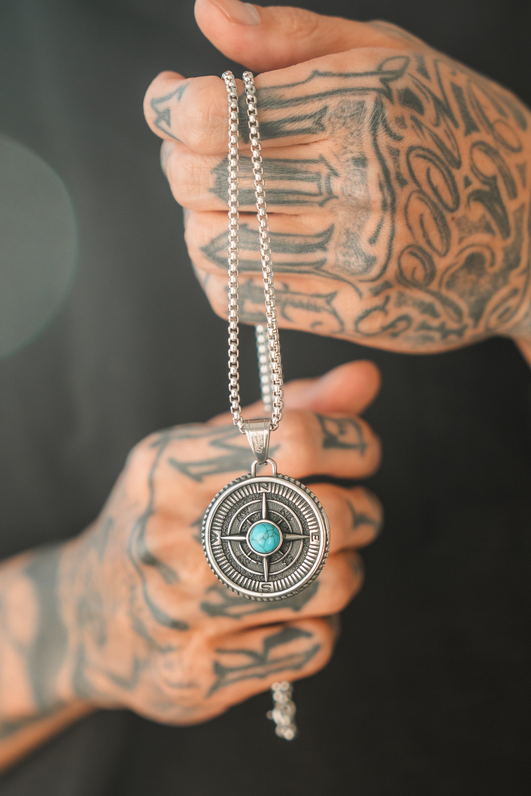 Tattooed hands display the Crossroads Necklace by Relentless Betrayal, a silver piece with a large round pendant. Featuring a central turquoise stone and intricate metalwork, it symbolizes lifes transitions and pivotal moments.