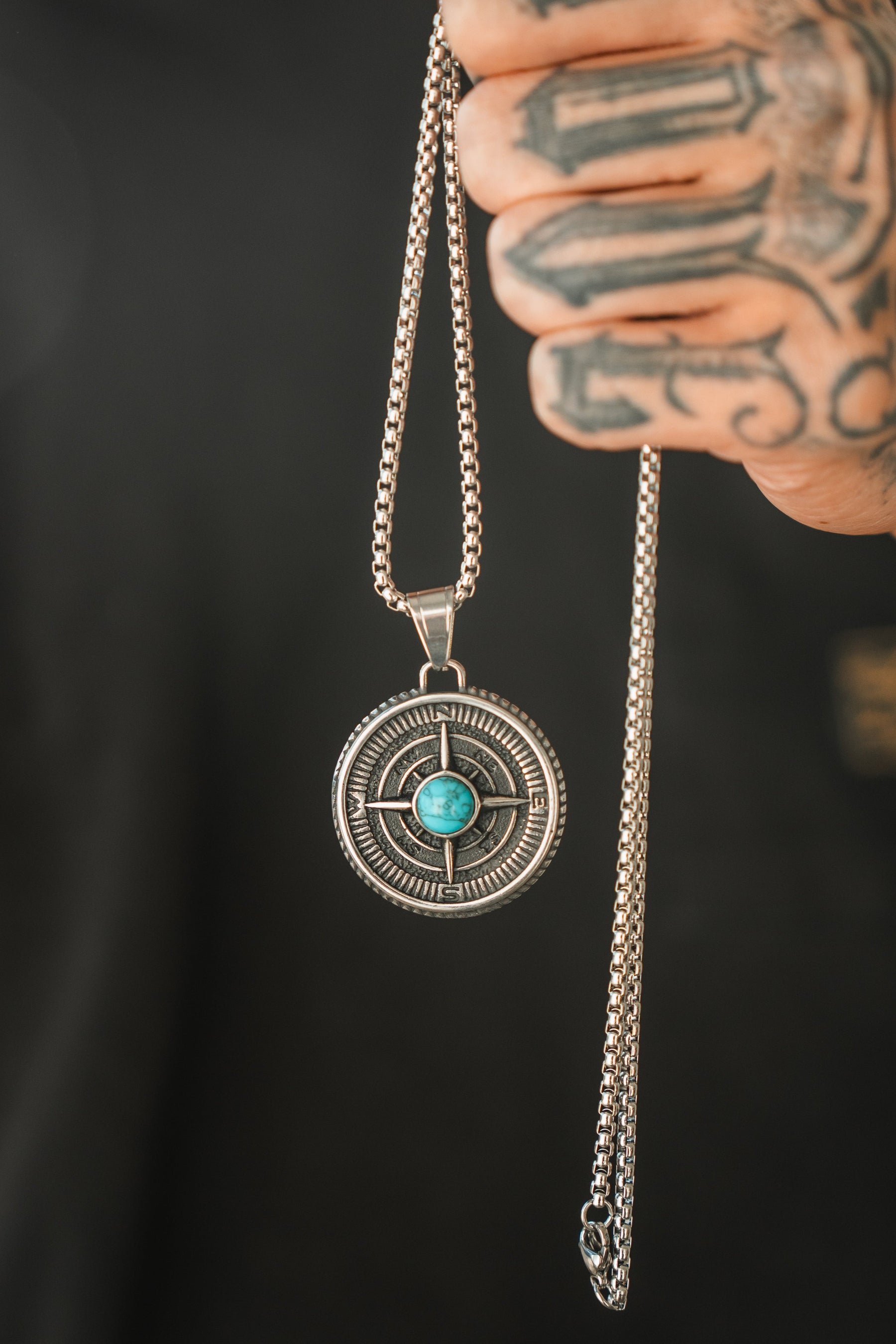 A tattooed hand holds a silver chain with the Crossroads Necklace by Relentless Betrayal, symbolizing transition. This round pendant features an ornate design centered with a turquoise stone, capturing pivotal moments against a dark background.