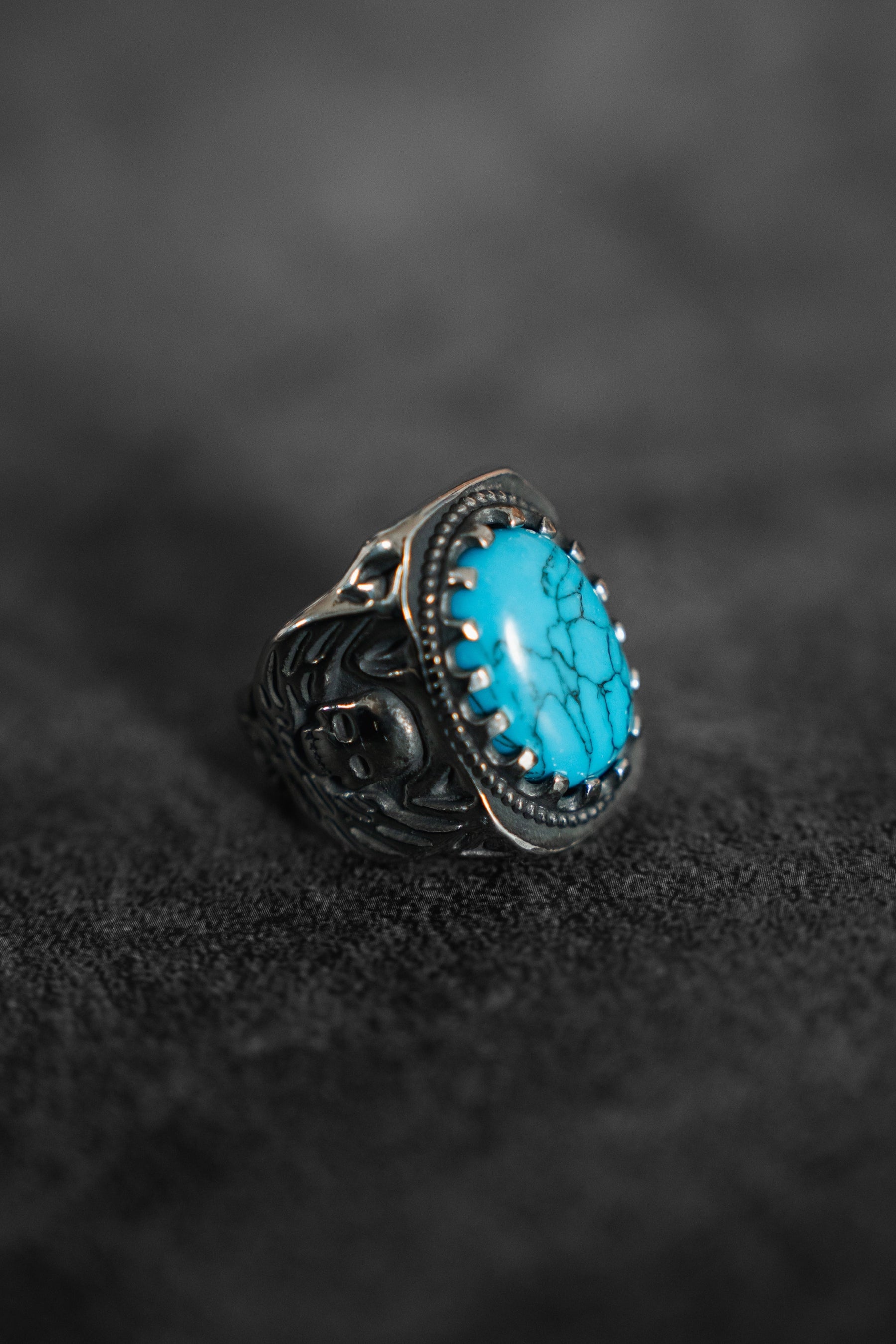 The Outsider Ring by Relentless Betrayal is a handcrafted silver piece with an intricate design, featuring a large oval turquoise stone in a prong setting, elegantly showcased against a textured dark gray fabric backdrop.